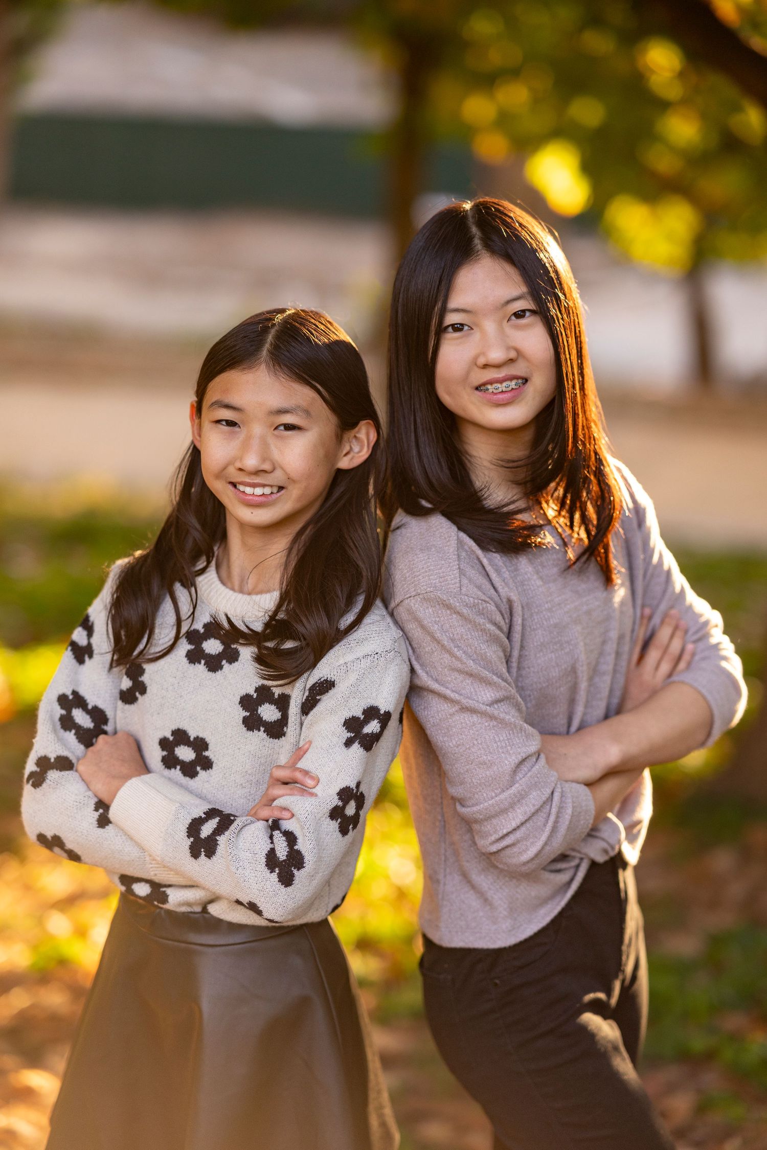 Two people in sweaters pose with crossed arms and bright smiles in outdoor autumn setting.