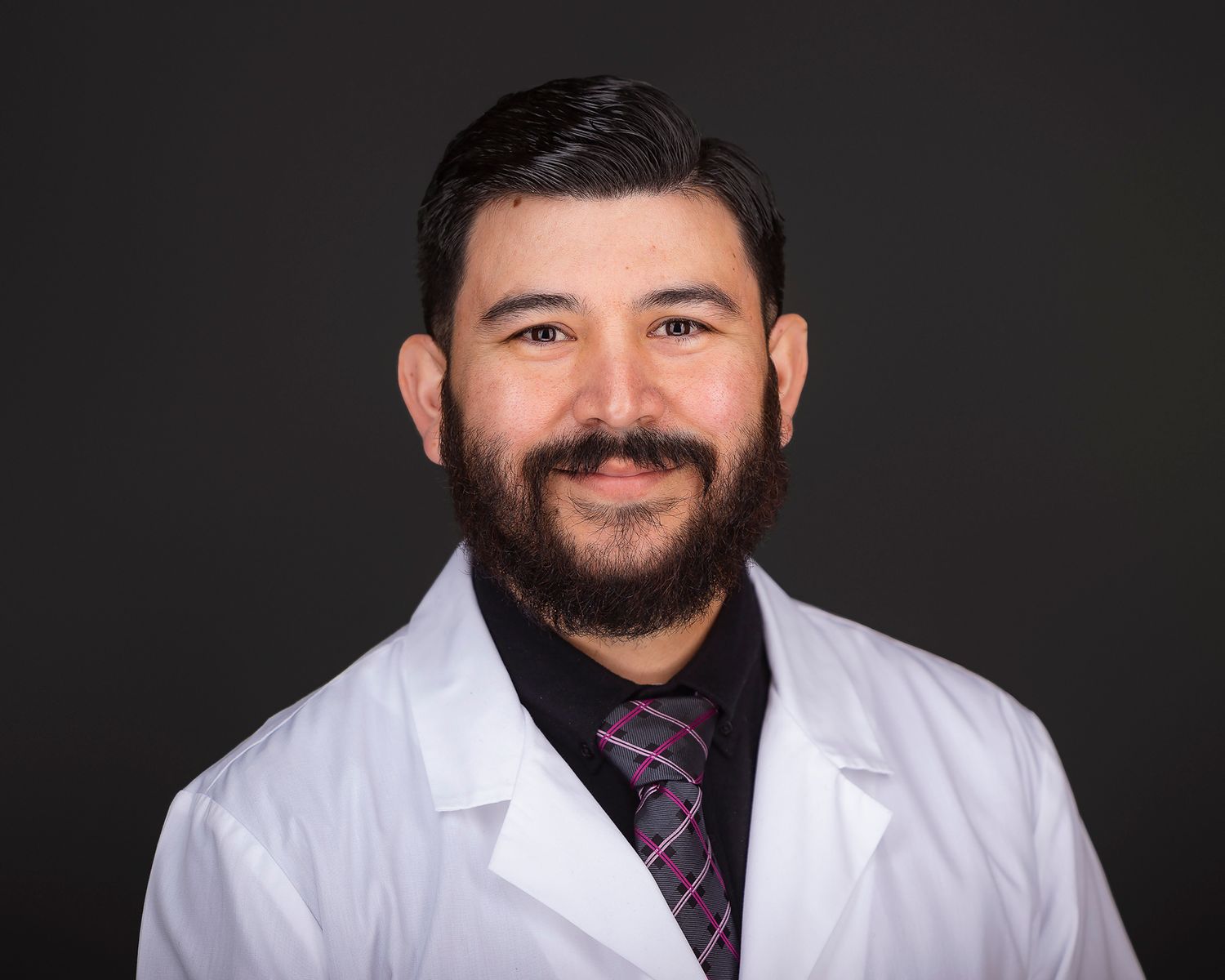 Professional headshot portrait of a healthcare provider wearing a white lab coat and purple striped tie against dark background.