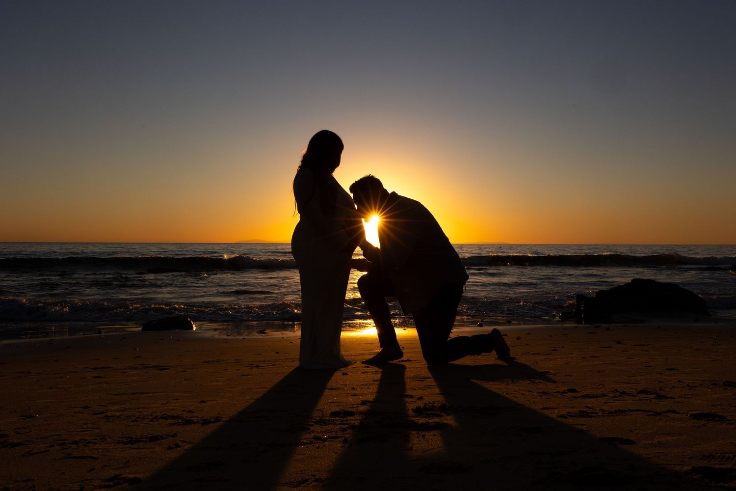 Silhouetted figures stand together on a beach at sunset creating romantic shadows against the golden sky in Huntington Beach.