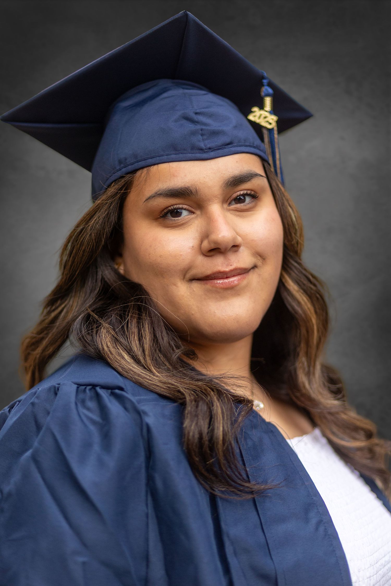 Young woman in navy blue graduation cap and gown smiling for a portrait photo against a gray background.