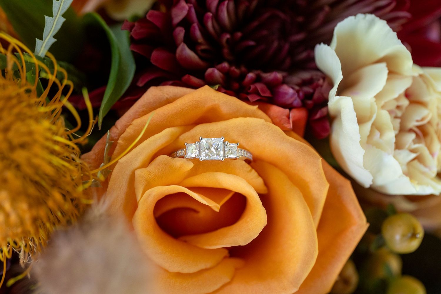 Close-up of orange and cream roses with wedding rings on textured background.