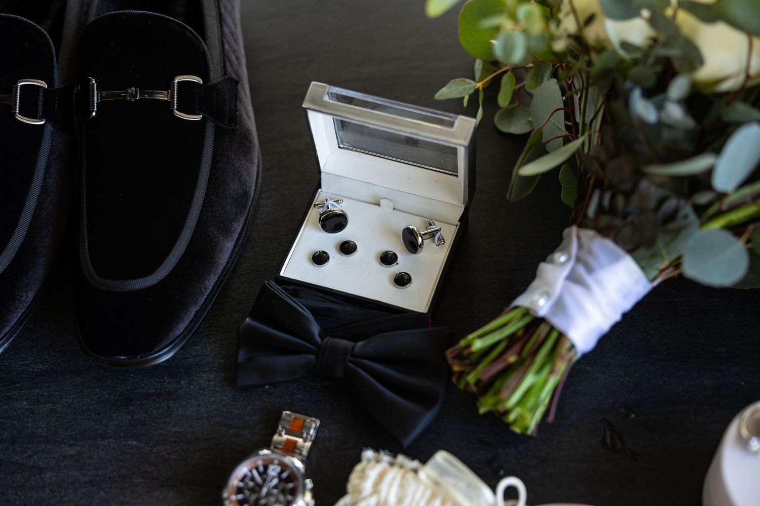 Wedding accessories including shoes, bouquet and cufflinks arranged on dark background.