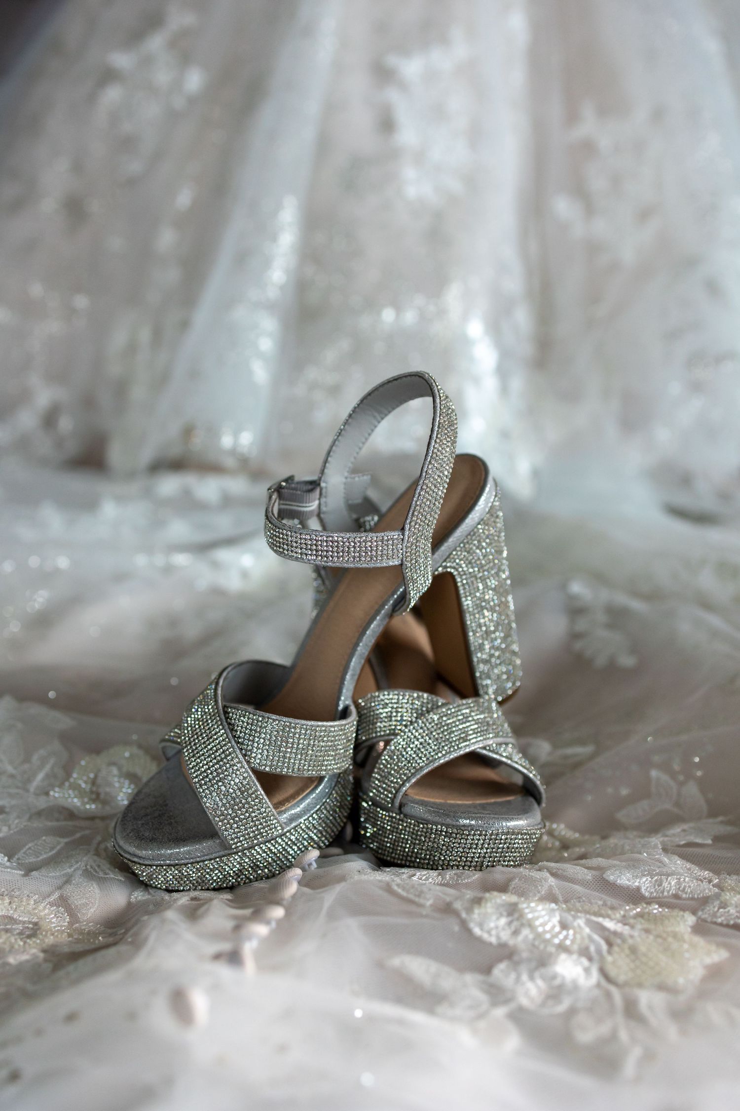 Sparkling silver wedding shoes with glitter and ankle straps laid out on white lace fabric.