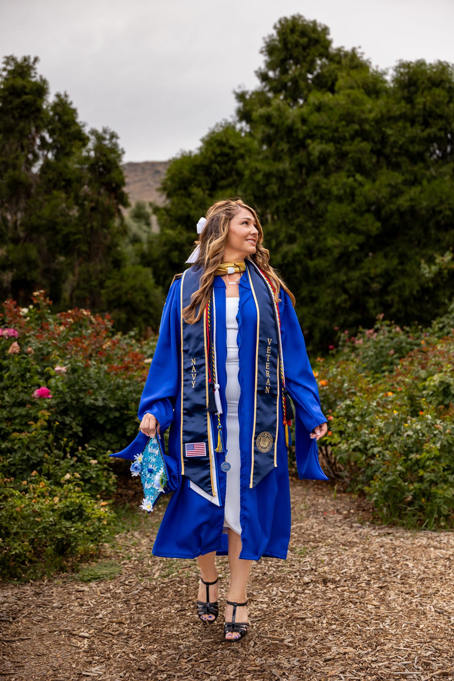 Graduate in royal blue commencement regalia posing in a rose garden.
