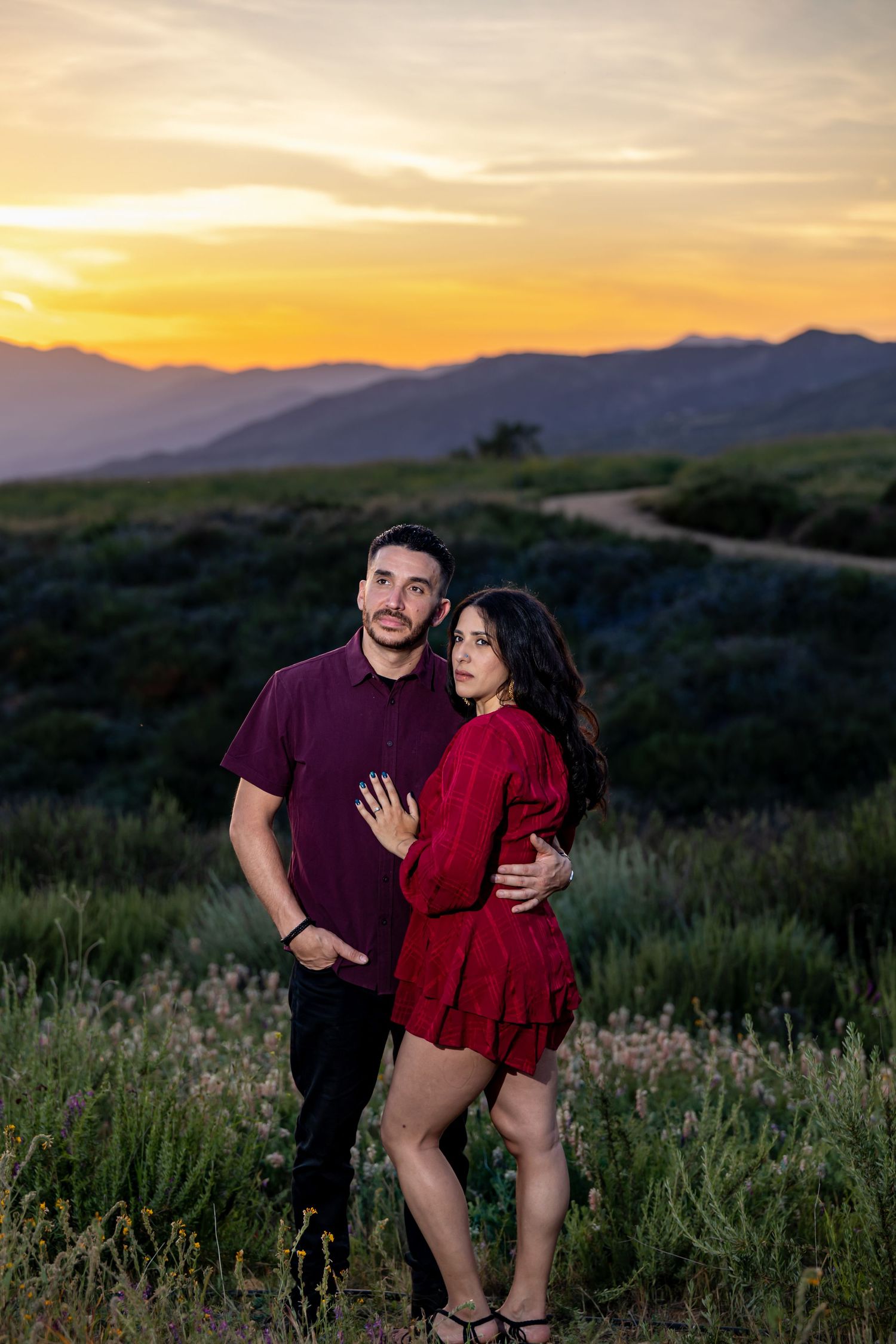 Happy couple in coordinating outfits posing in field with dramatic sunset sky behind them.