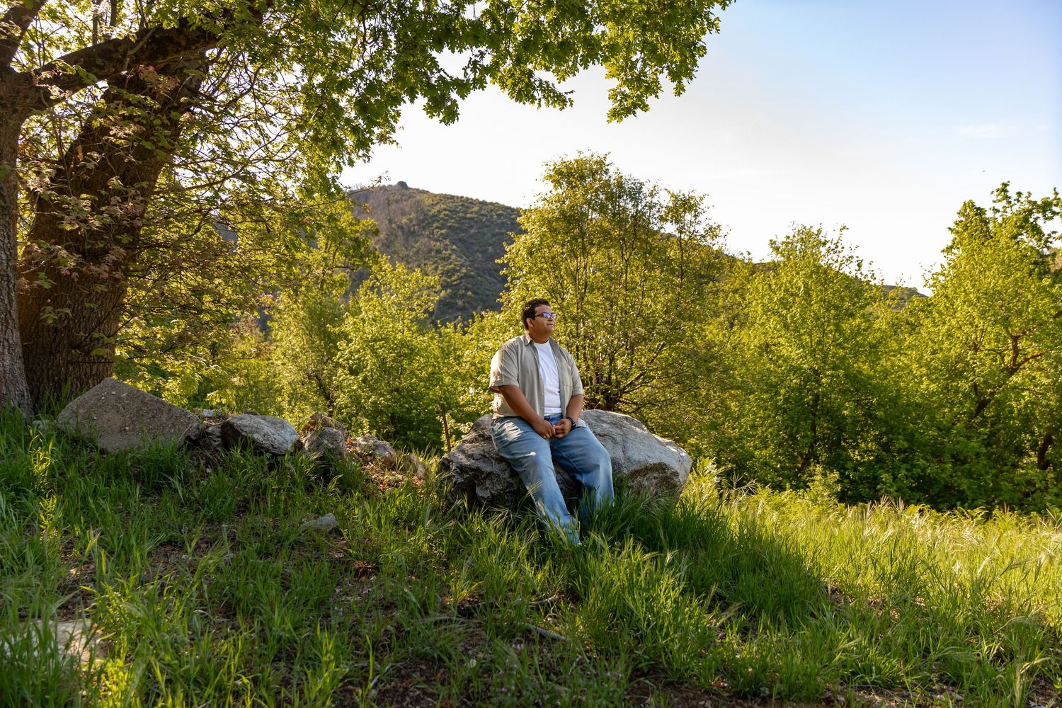A person sits relaxing on a grassy hillside at sunset with trees and mountains in the background in Yucaipa.