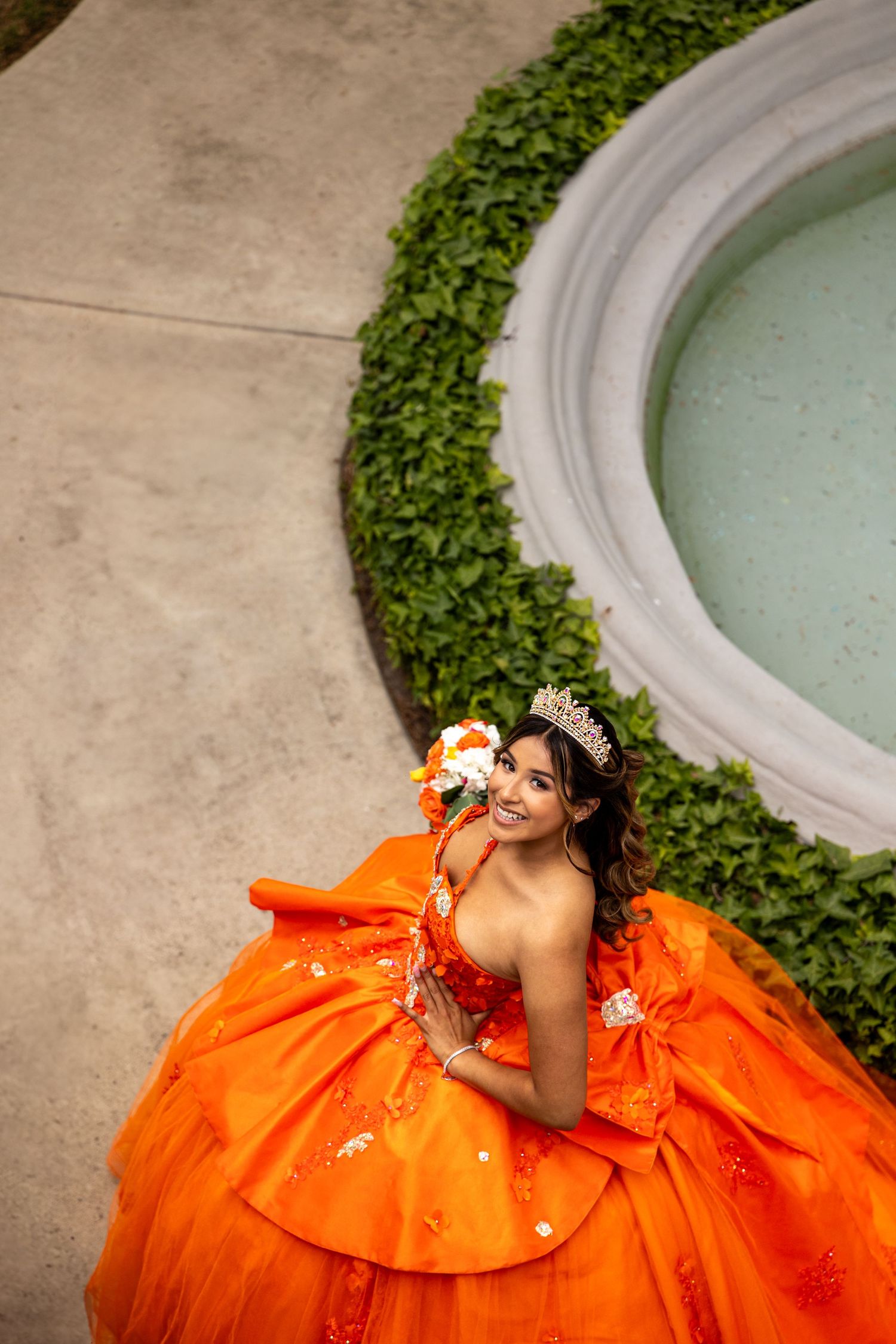 A vibrant orange quinceañera dress displayed near a curved pool edge with greenery.