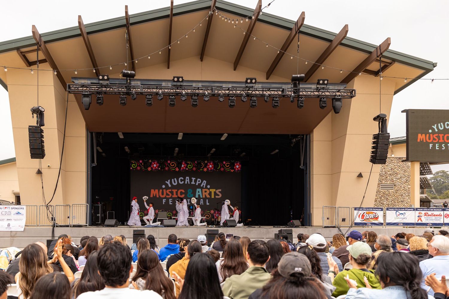 People gather in front of an outdoor stage at a festival or concert venue with beige walls and a covered roof area.