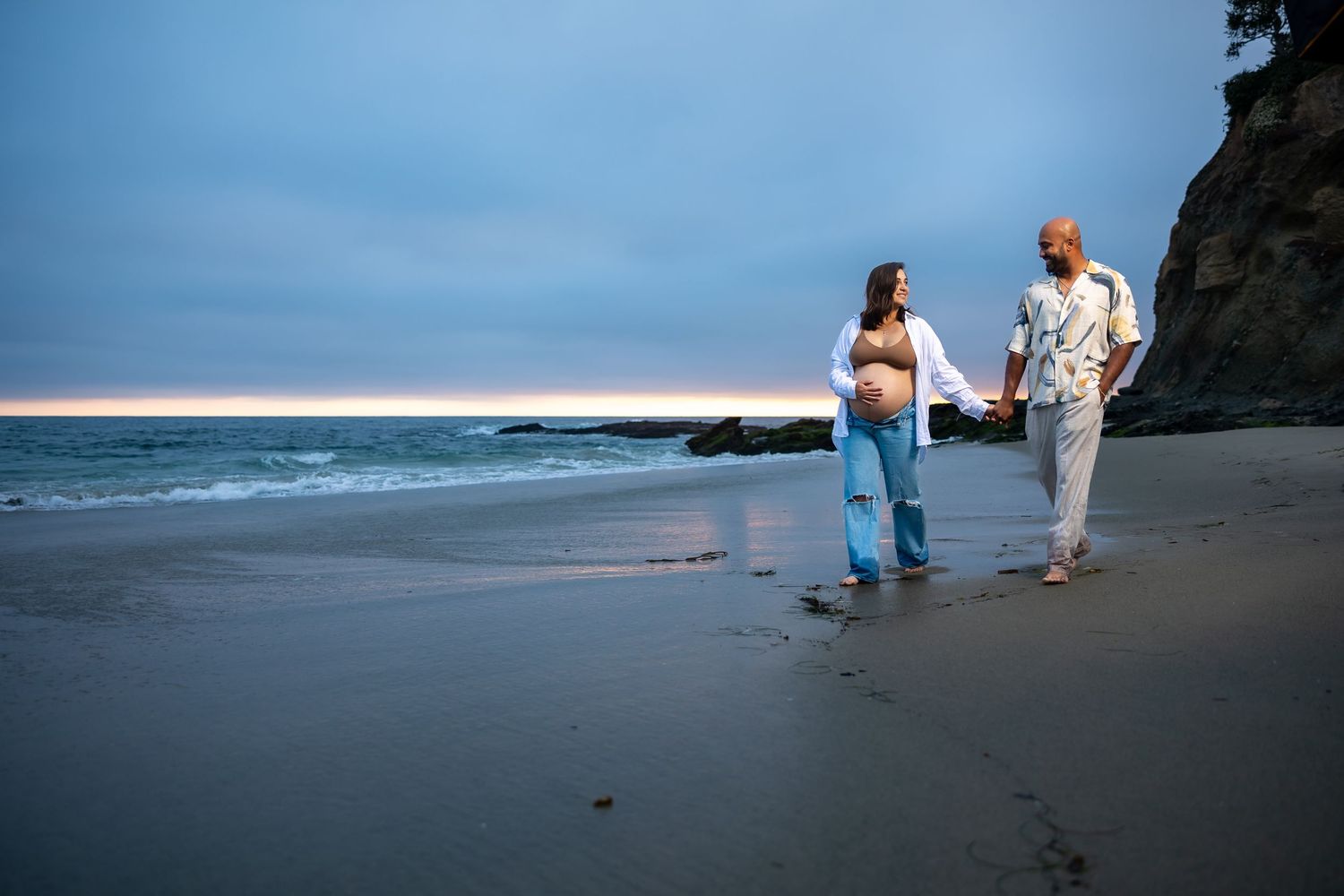Silhouettes of beachgoers stroll along a sandy shore at sunset with blue ocean and cloudy sky in the background in Huntington Beach.