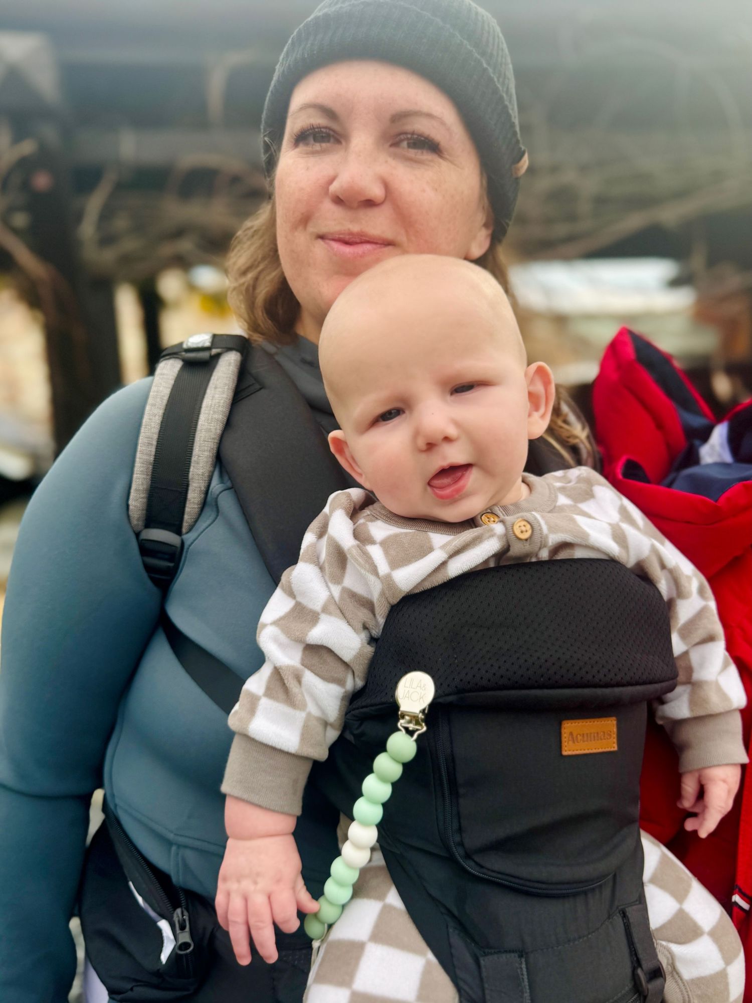 Winter scene with baby in patterned outfit being carried in black baby carrier.