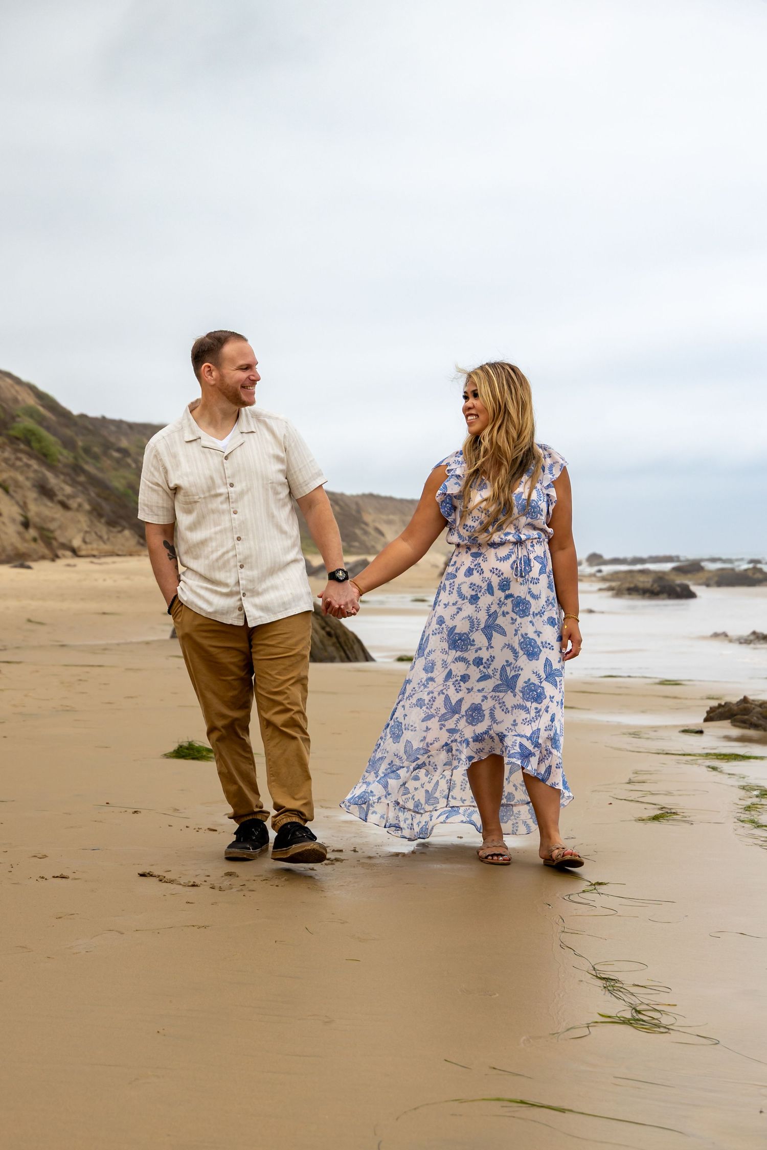 Couple walking hand in hand on beach wearing coordinated outfits with blue floral dress and khaki.