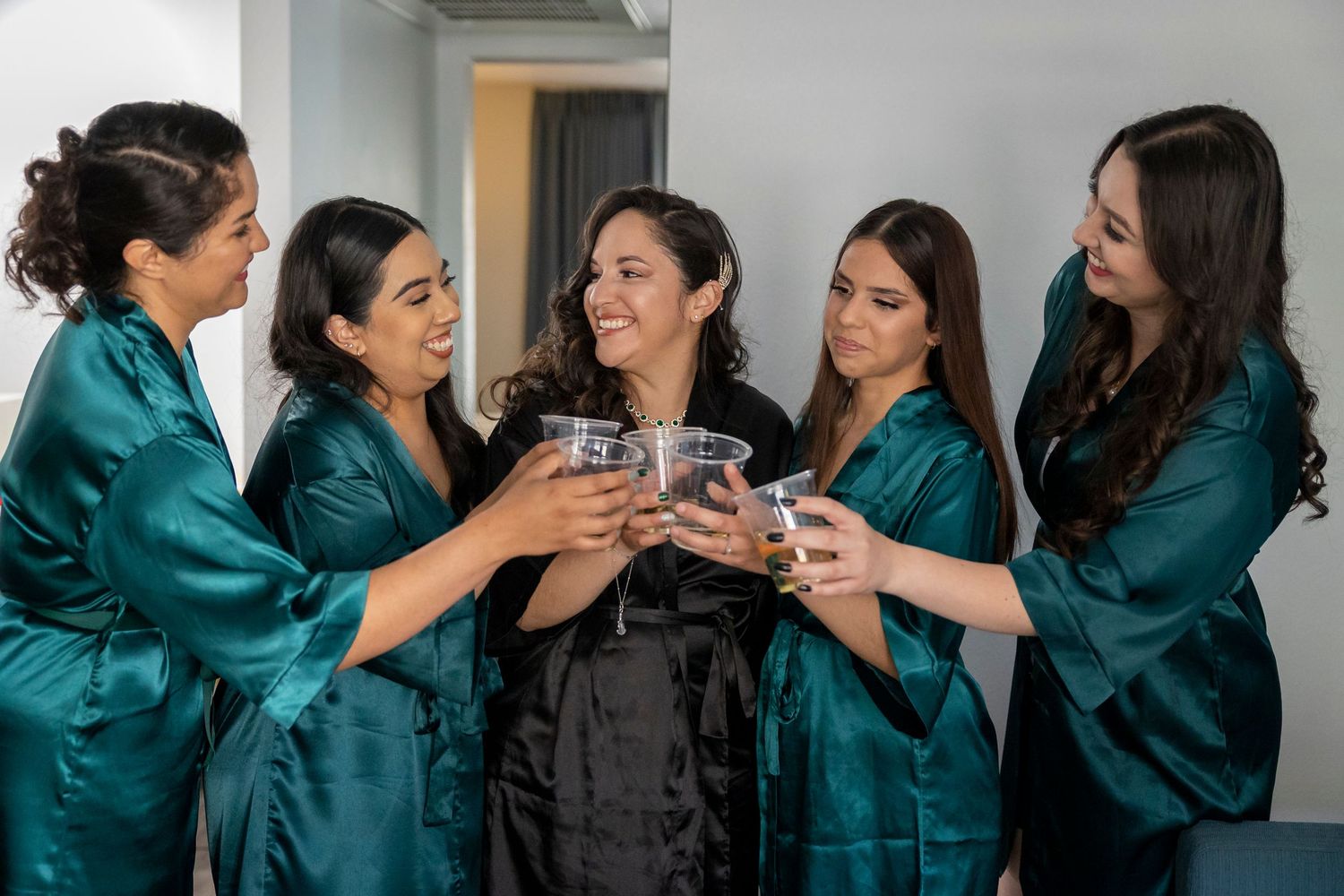 Bridal party in teal satin robes toasting with champagne glasses during wedding preparations.