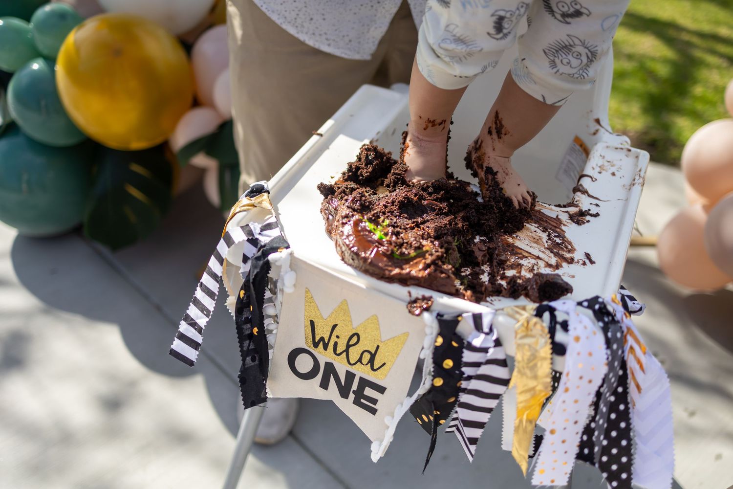 Small hands smashing chocolate cake with Wild One banner and balloon decorations.
