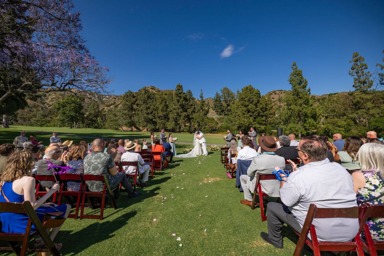 Outdoor wedding ceremony setup with red chairs on a green lawn surrounded by trees under bright blue sky in Burbank