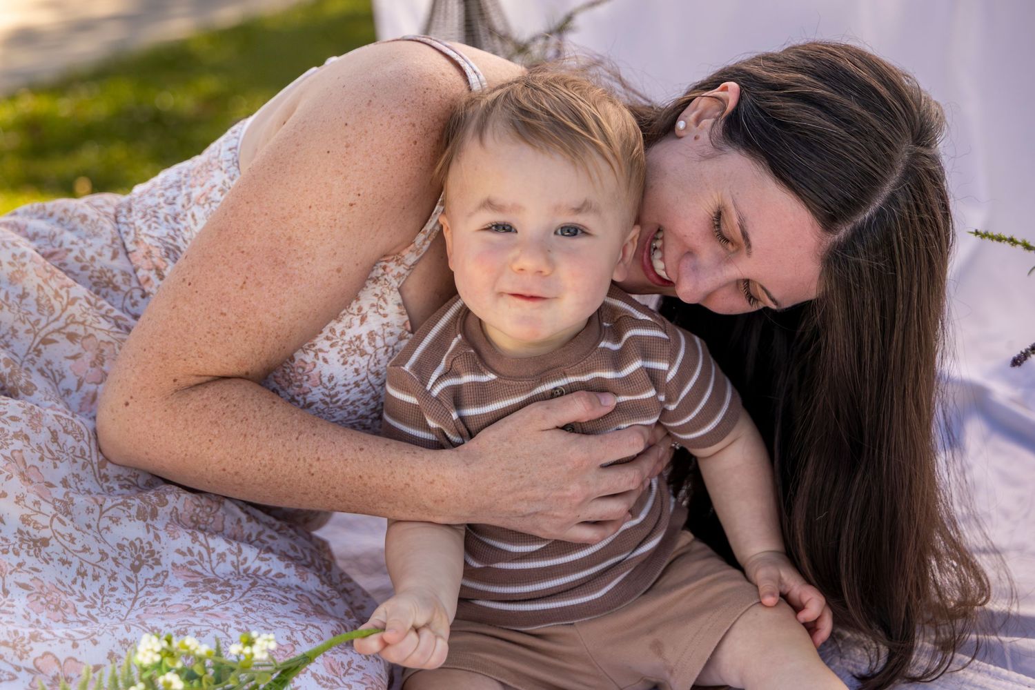 A mother tenderly embraces her smiling baby boy outdoors on a soft blanket in warm natural light.