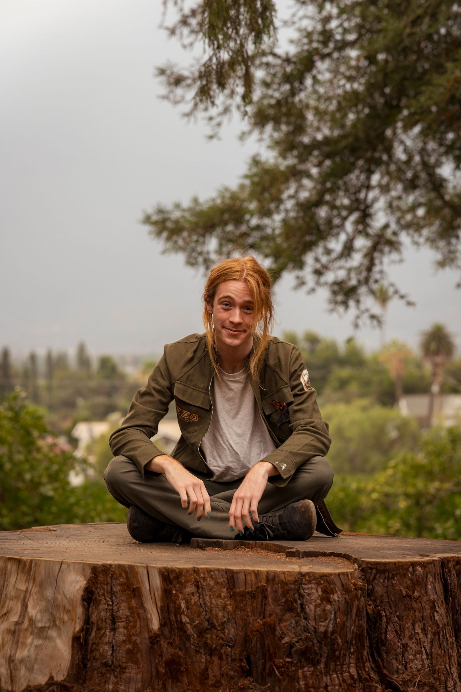 Casual portrait on a large tree stump wearing an olive jacket and dark pants in Redlands.