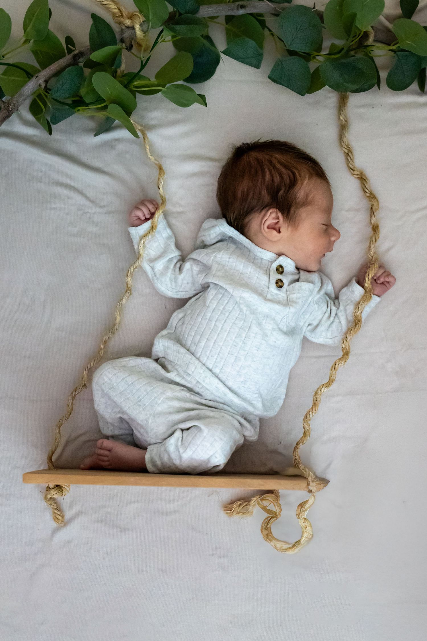 Baby in white outfit lying on swing with greenery decoration.