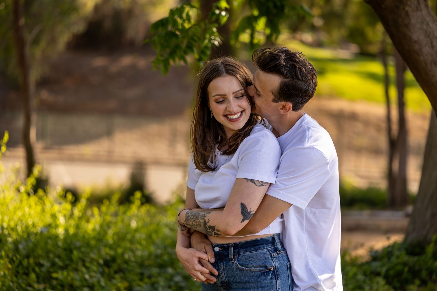 A romantic couple shares a joyful embrace under green trees in a sunny outdoor setting.