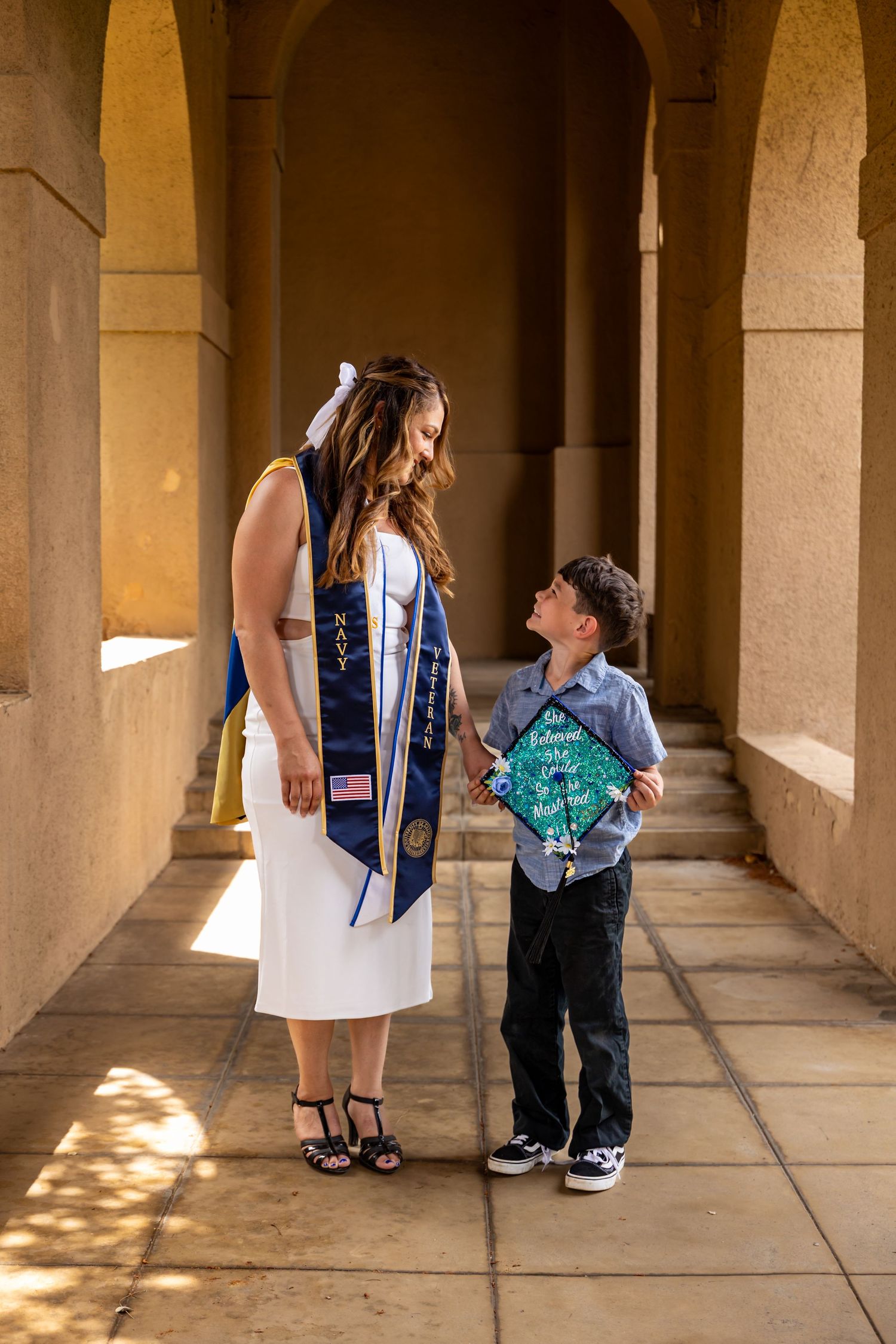 Graduate and young companion walking through sunlit stone archway in Riverside.