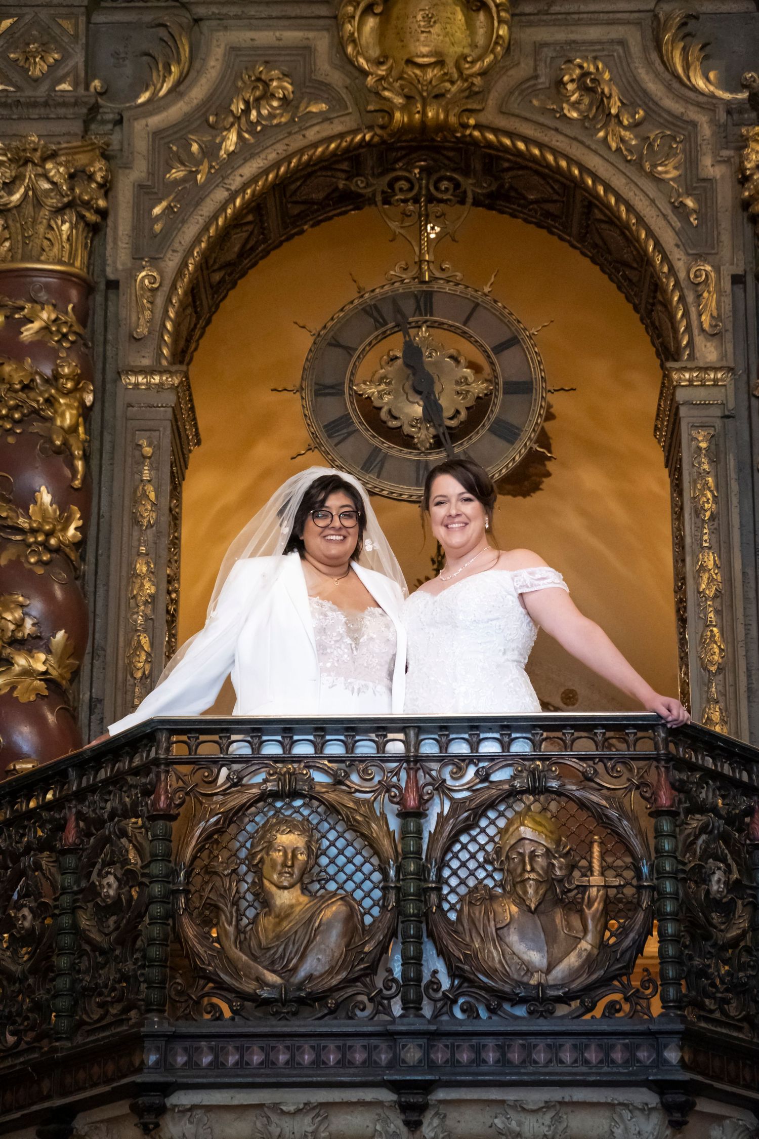 Two brides pose together on an ornate balcony with a vintage clock behind them.