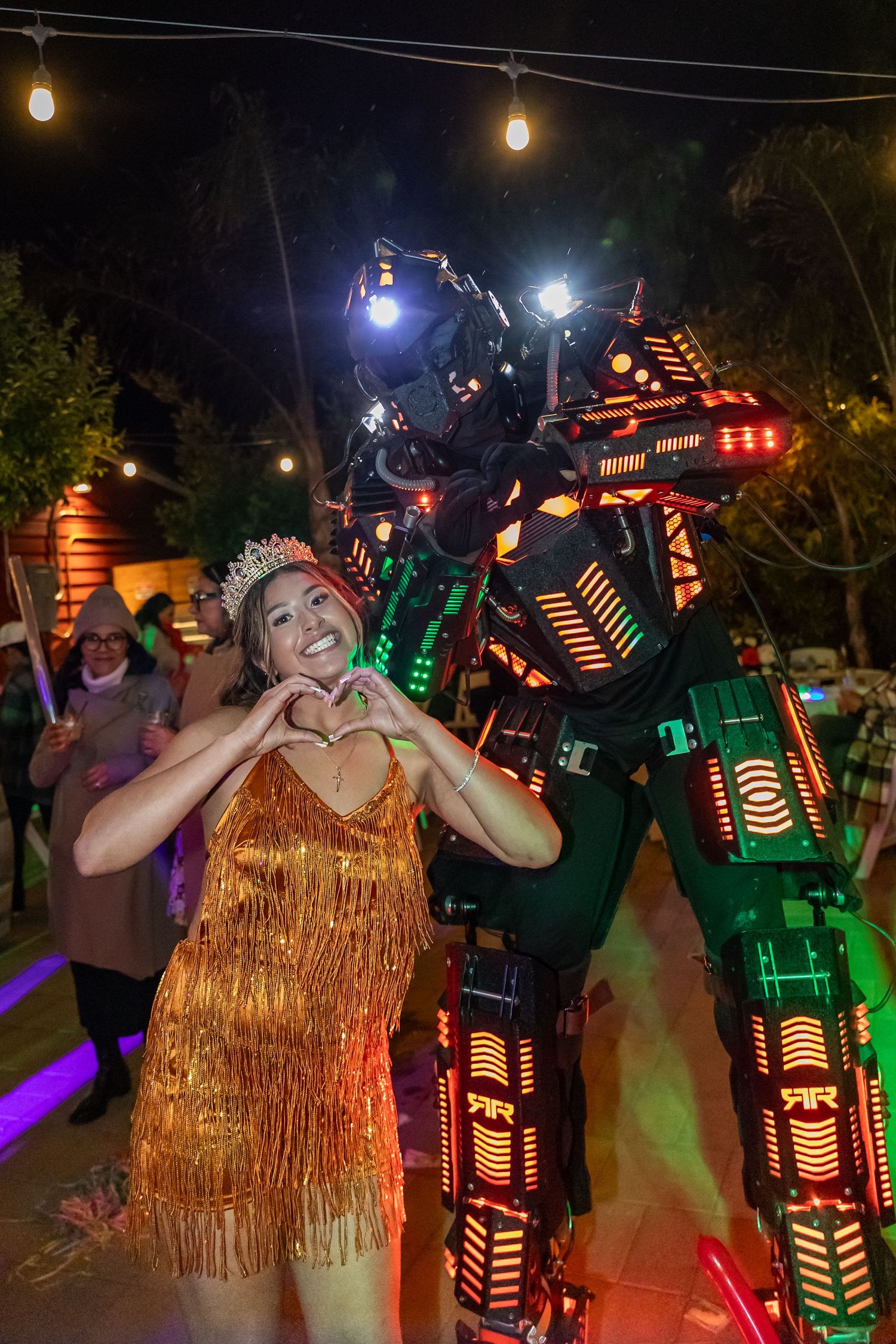 Person in gold fringe dress posing with LED robot costume performer at night event.