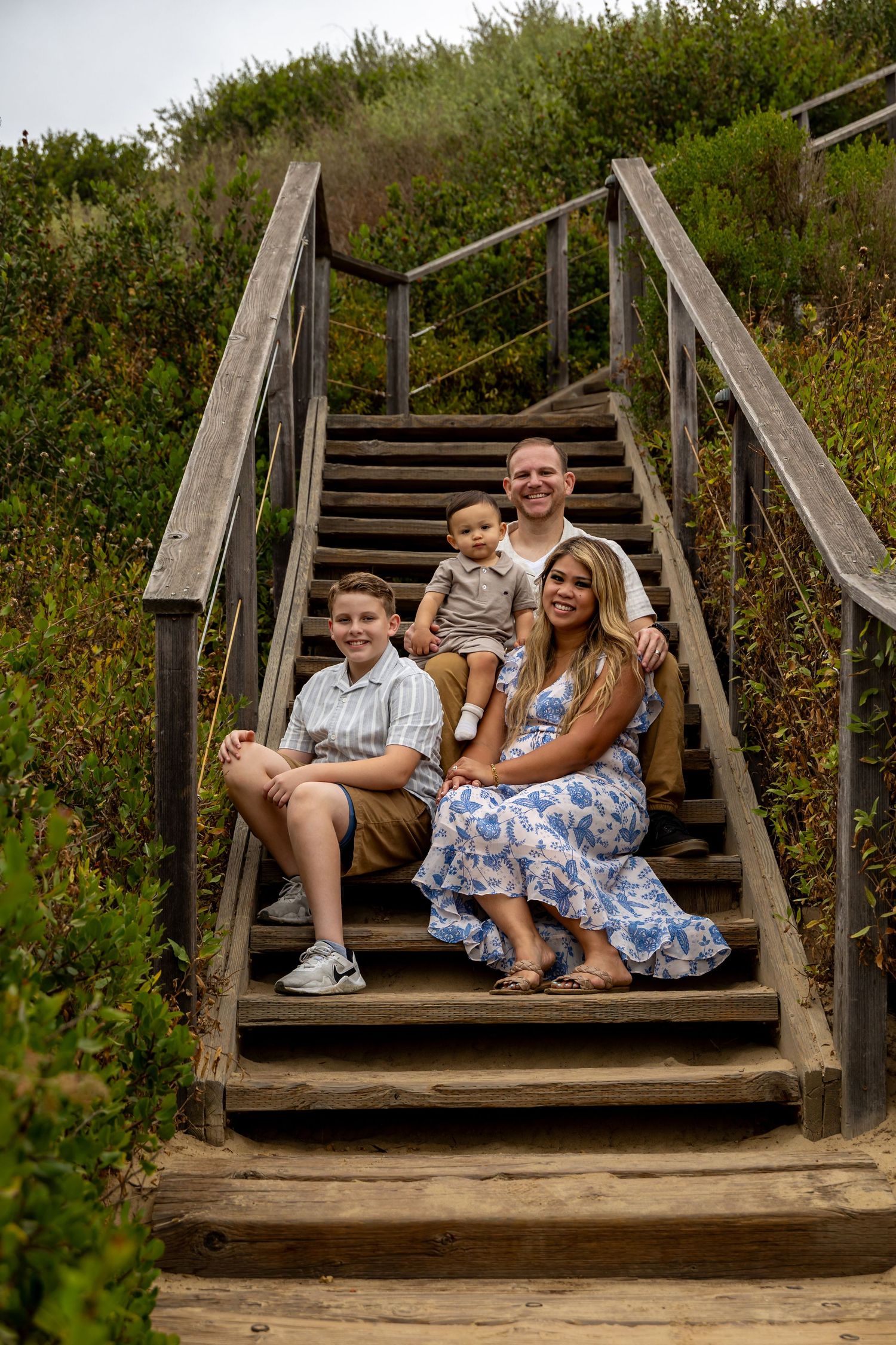 Family sitting together on wooden beach stairs surrounded by green coastal vegetation.
