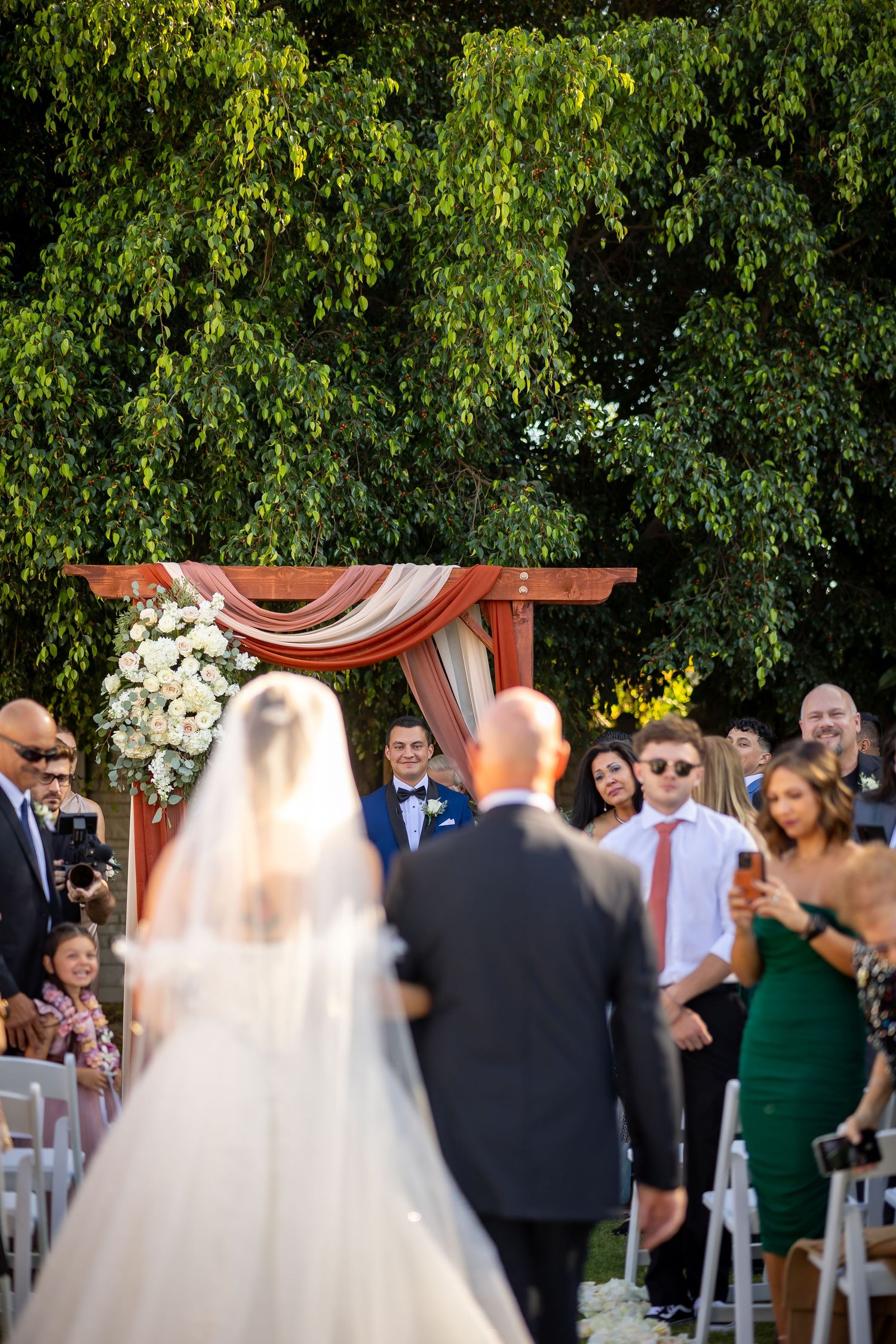 An outdoor wedding ceremony under lush green trees with a decorated wooden arch.
