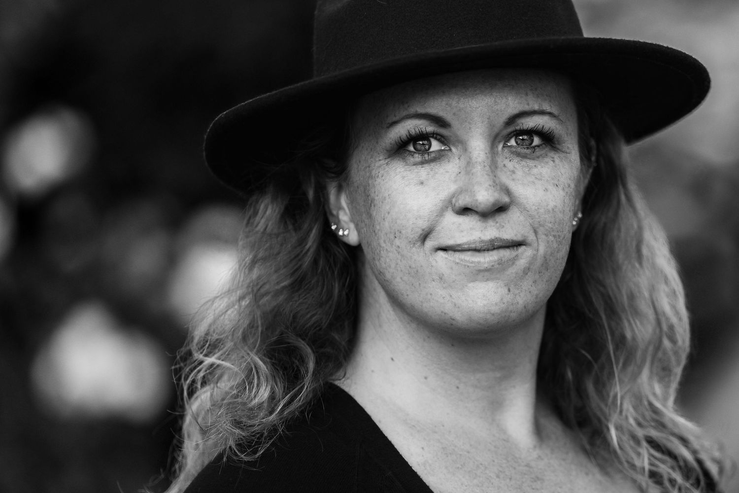 Black and white portrait of a smiling woman with curly hair wearing a wide-brim hat outdoors.