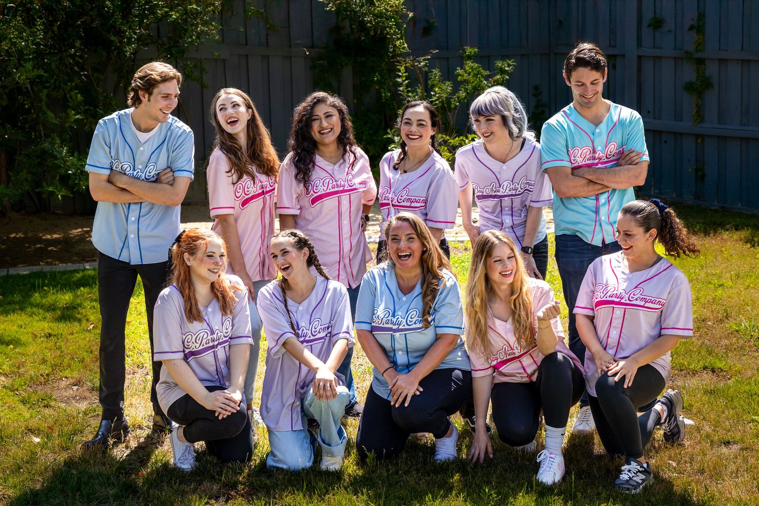 Group of young people in coordinated pink and blue outfits posing together on sunny grass for a casual outdoor photo.