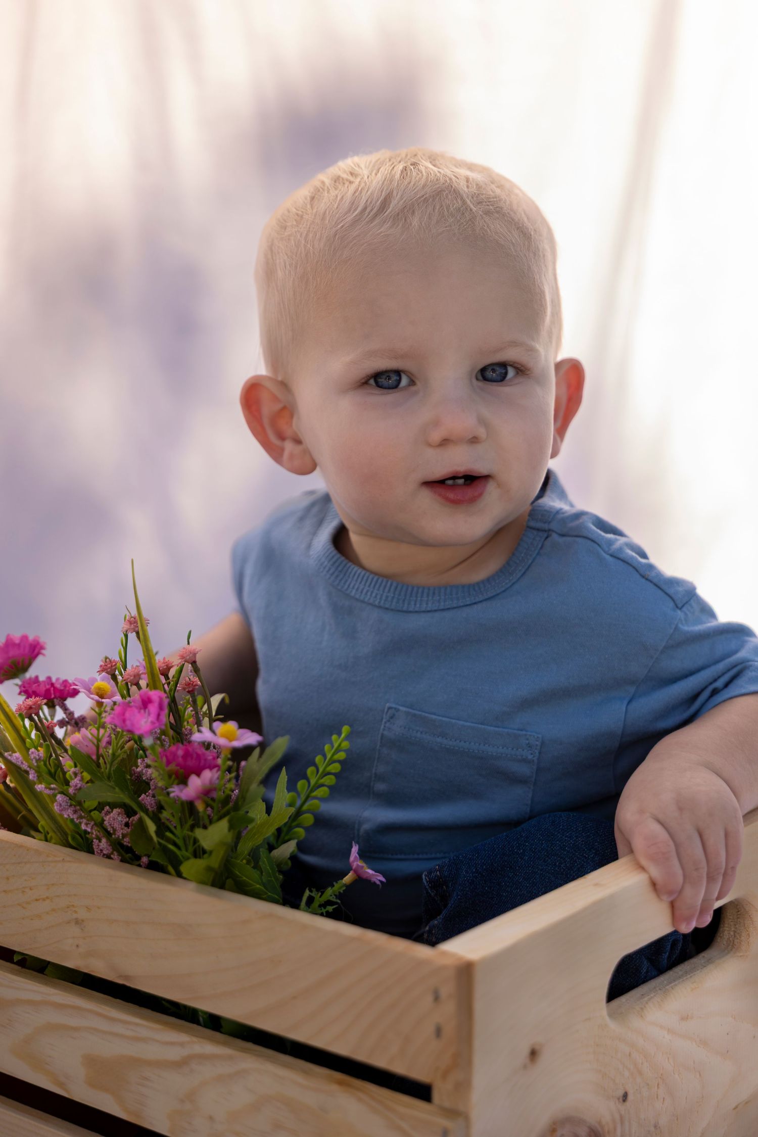Toddler boy in blue shirt sitting in wooden crate with pink flowers against soft purple backdrop.