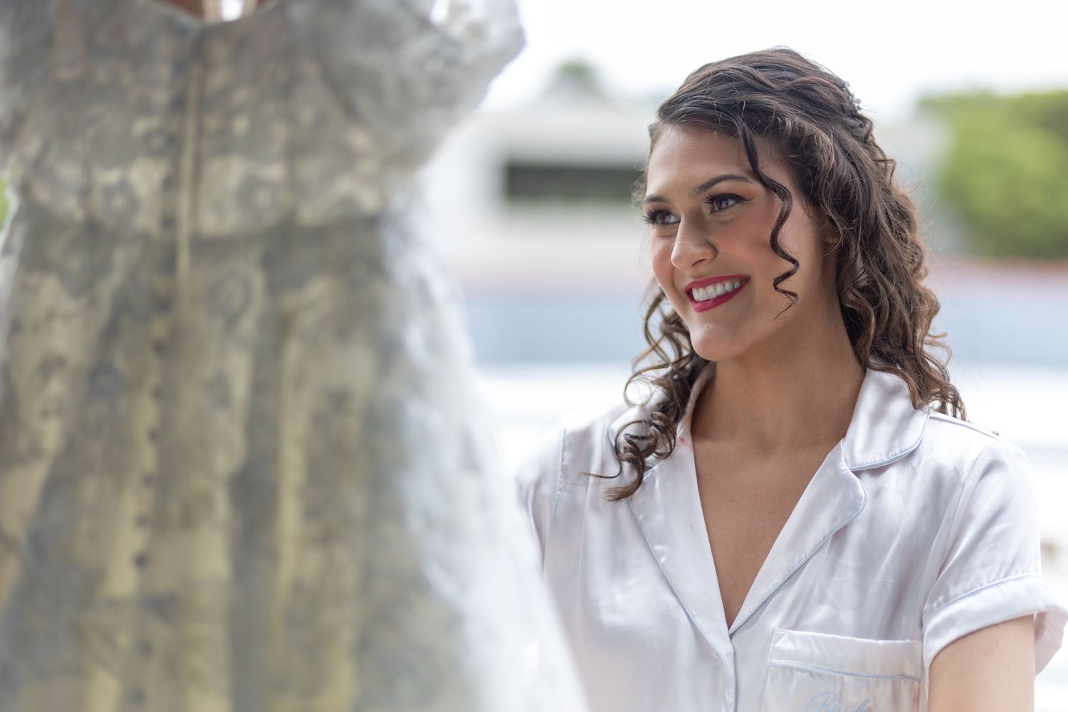 A bride in a white button-up shirt smiles while adoring her wedding dress in Huntington Beach