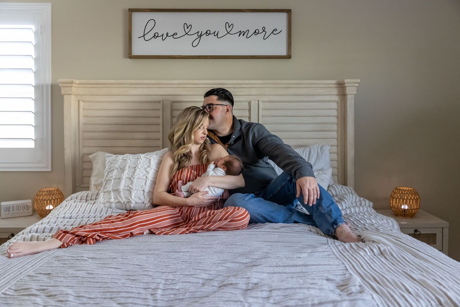 Couple cuddling with newborn baby on bed beneath rustic wall art.