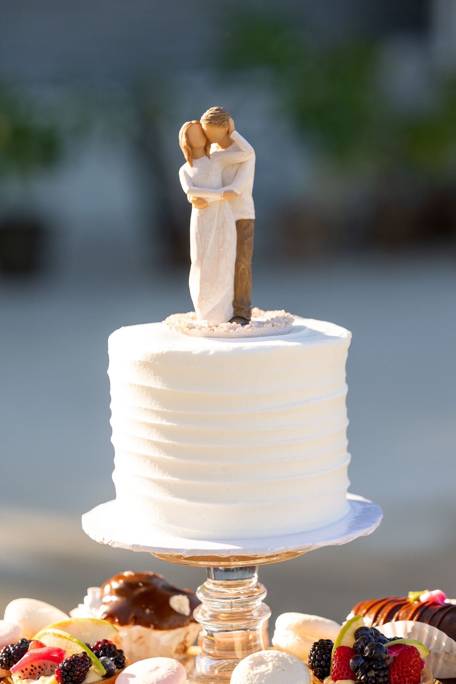 An elegant white wedding cake with multiple tiers sits atop a glass stand with fresh berries and floral decorations.