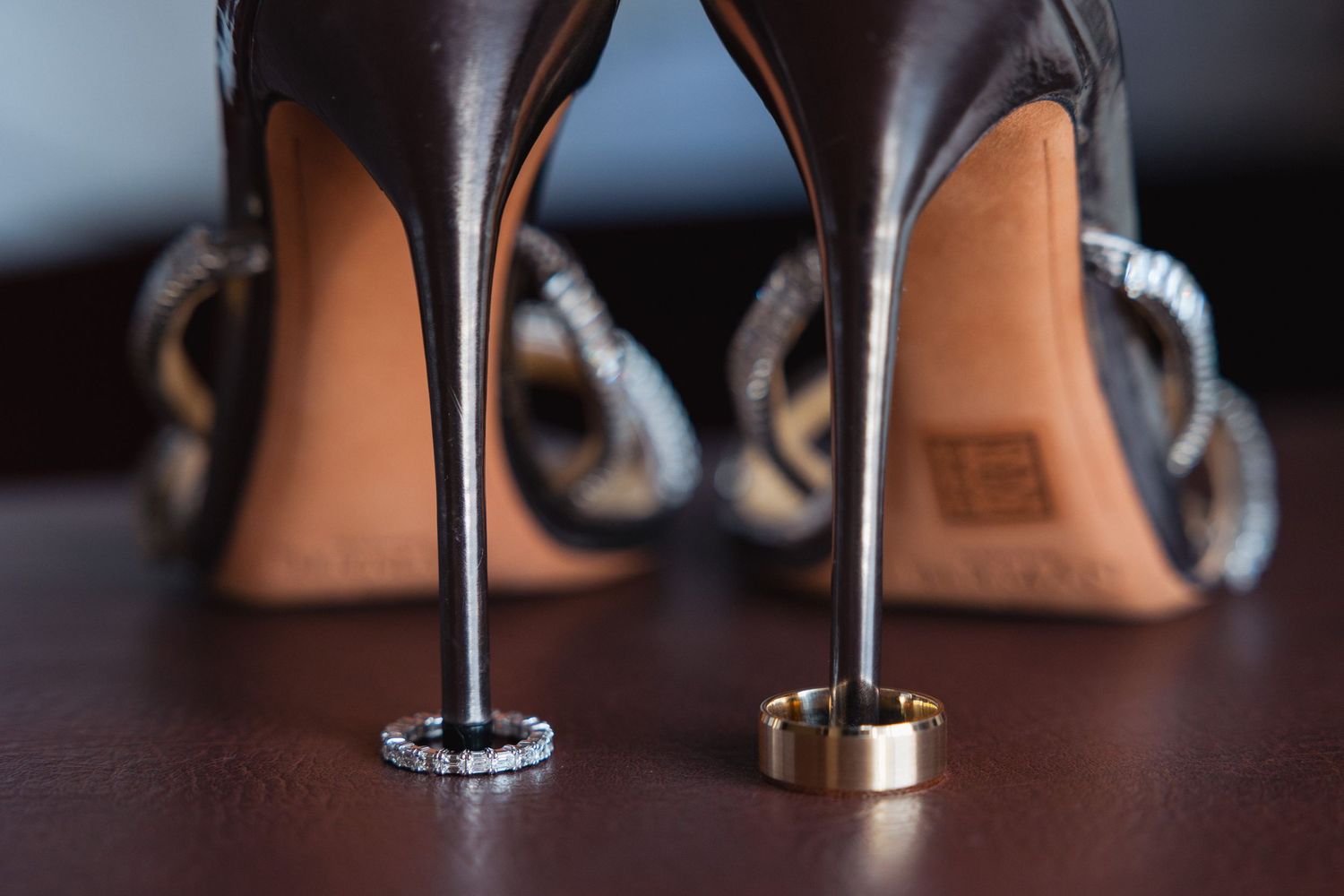 Wedding rings and bands photographed between elegant high heels on a dark surface.
