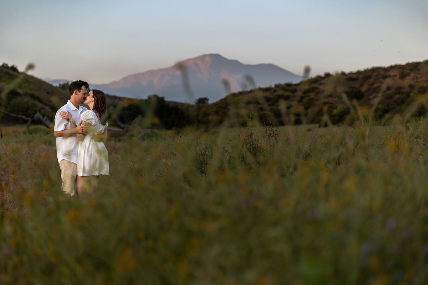 Two figures embrace in a grassy field with mountains and sunset in the background.