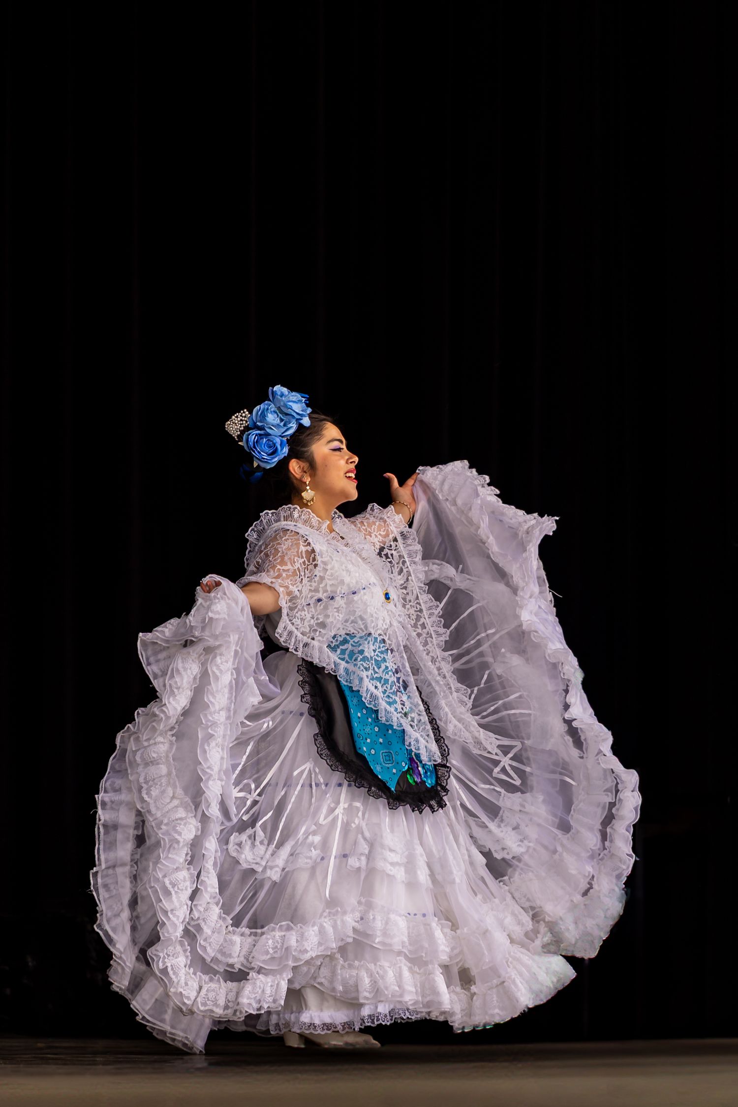 Four sequential photos show a dancer in a flowing white lace dress with blue accents performing a twirling movement on stage.