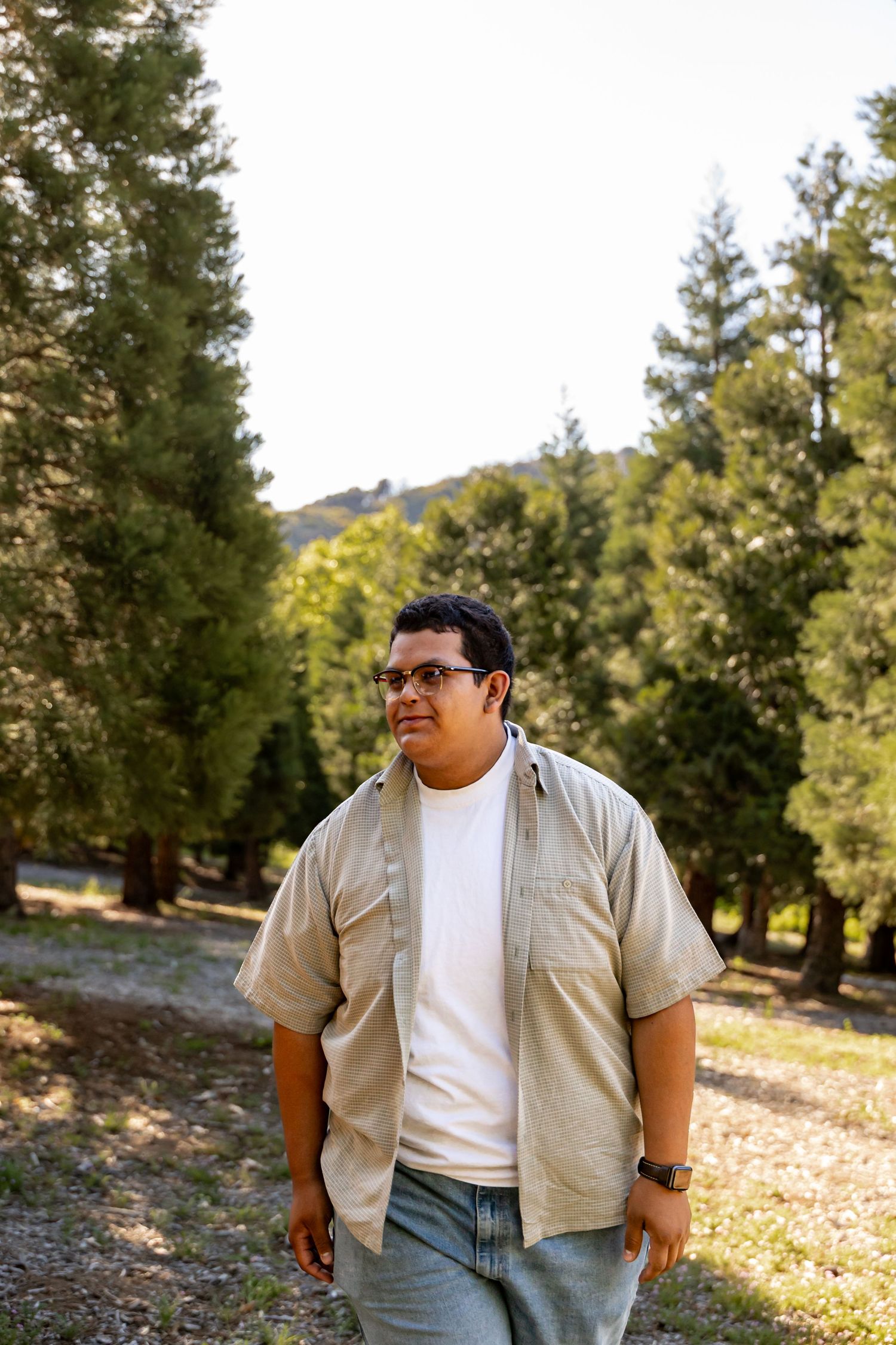 Someone wearing glasses, a beige shirt and white t-shirt poses casually in a scenic forest setting at golden hour in Yucaipa.