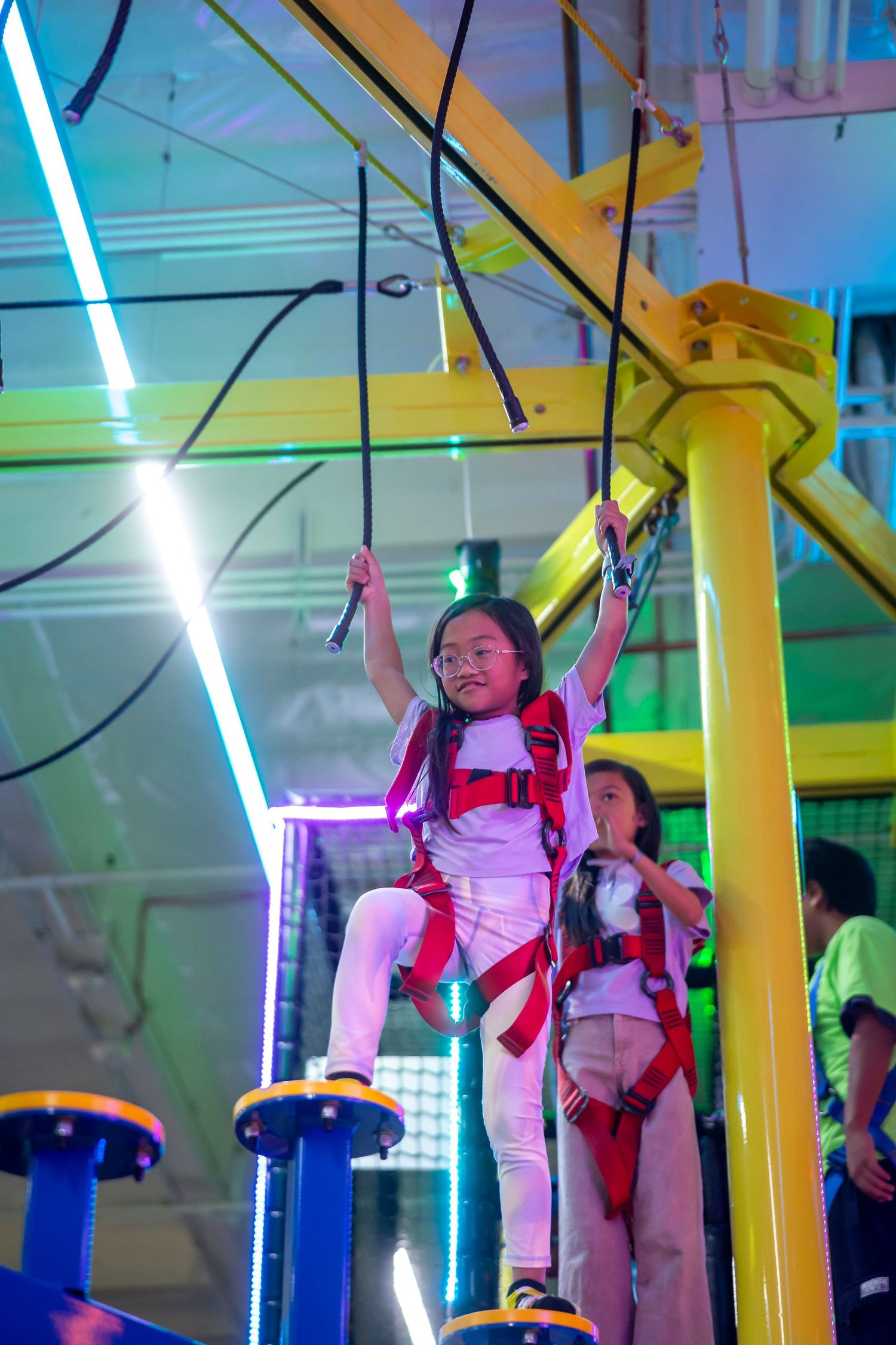 Kids climbing and swinging on yellow ropes in a neon-lit indoor playground.