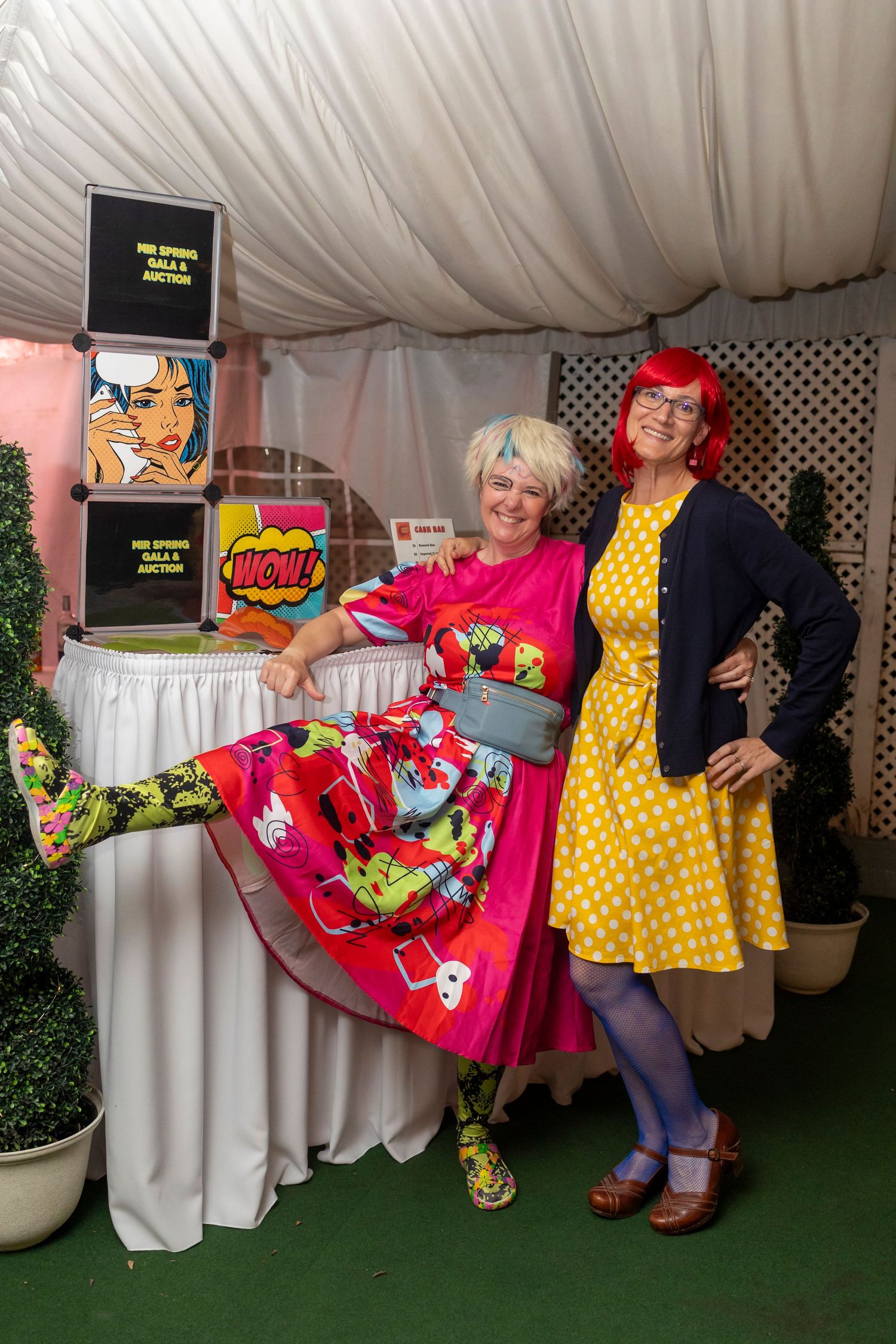 Group wearing colorful retro-style dresses posing against a white backdrop with greenery.
