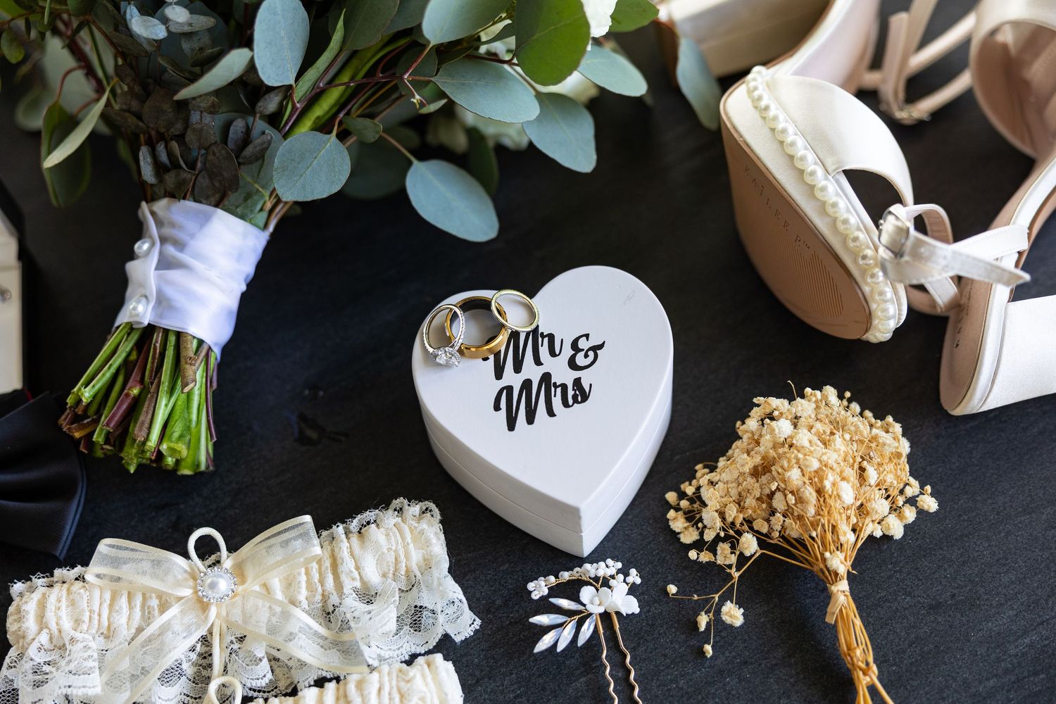 Wedding details including a bouquet, heart-shaped Mr & Mrs sign, and accessories arranged on dark surface.