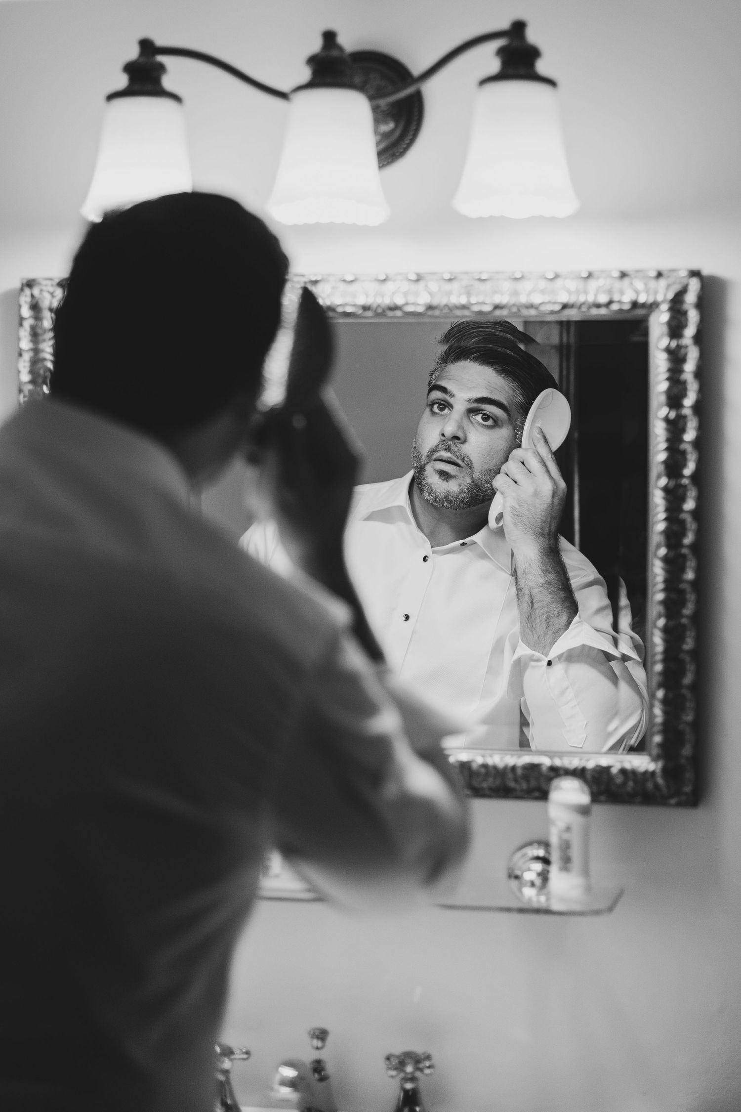 Black and white reflection of groom getting ready in bathroom mirror.