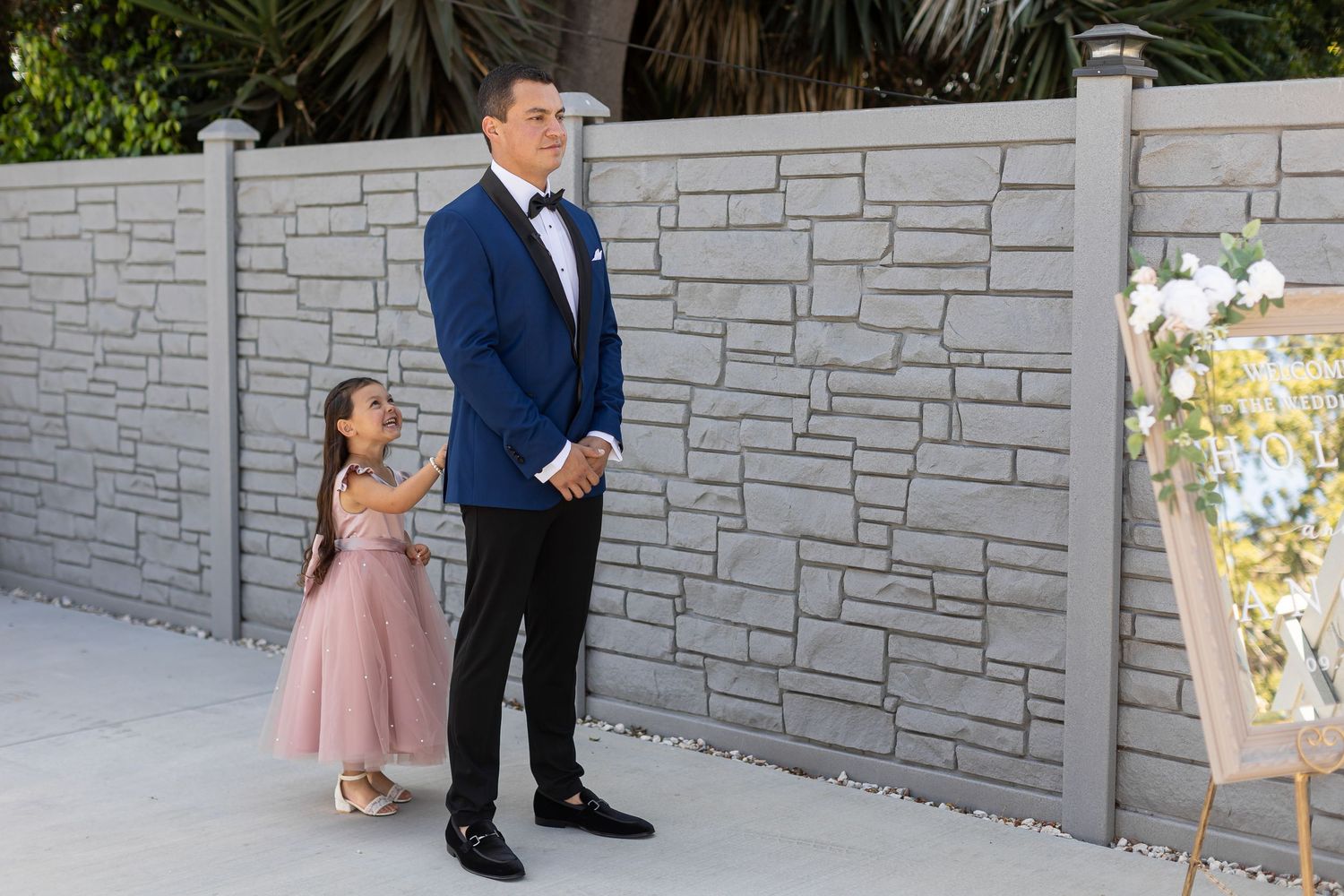 A pair in formal attire stand near a modern stone wall during a wedding celebration.
