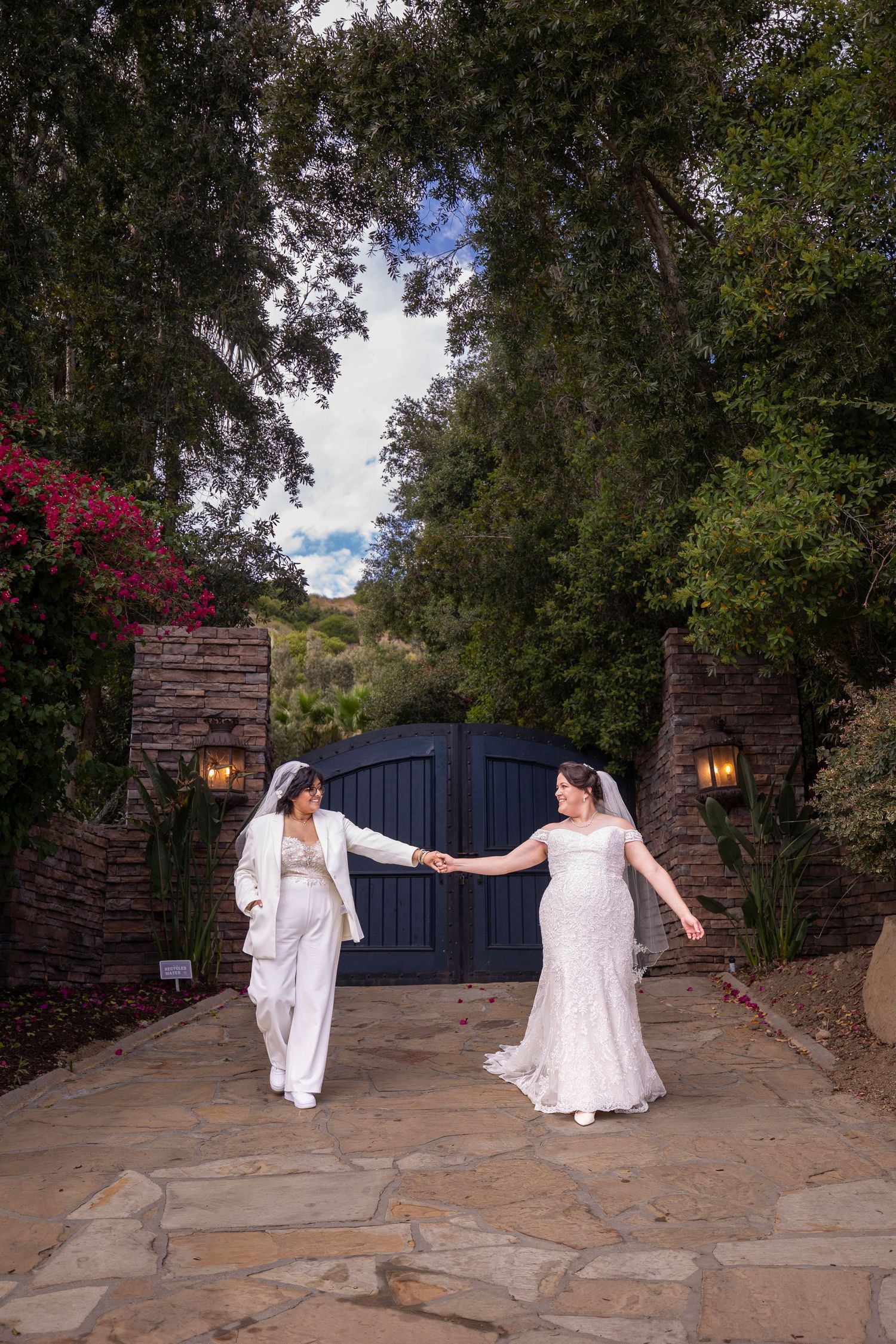 Two people in white dresses dancing on a stone patio with flowering trees and rustic walls.