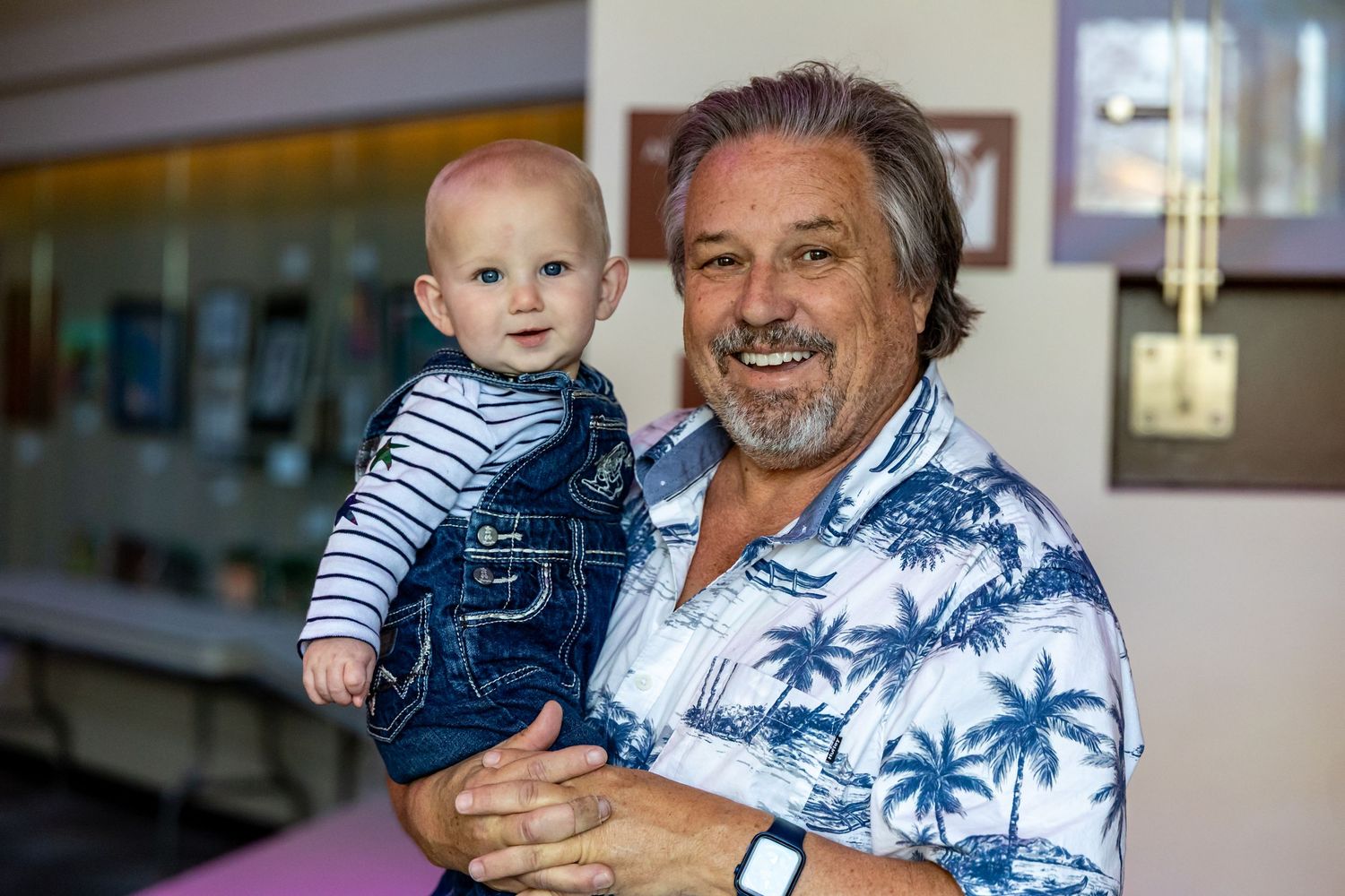 Adult wearing tropical shirt holds small child in striped overalls at indoor event.