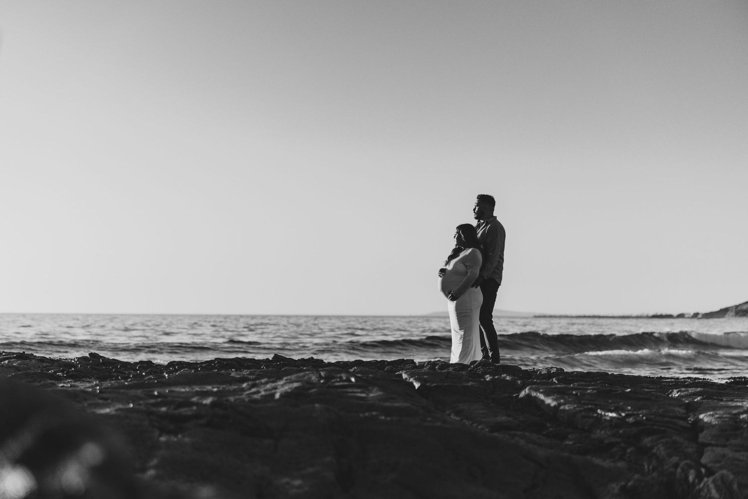 Silhouette of two people embracing on rocky shoreline at sunset with ocean waves in black and white in Huntington Beach.