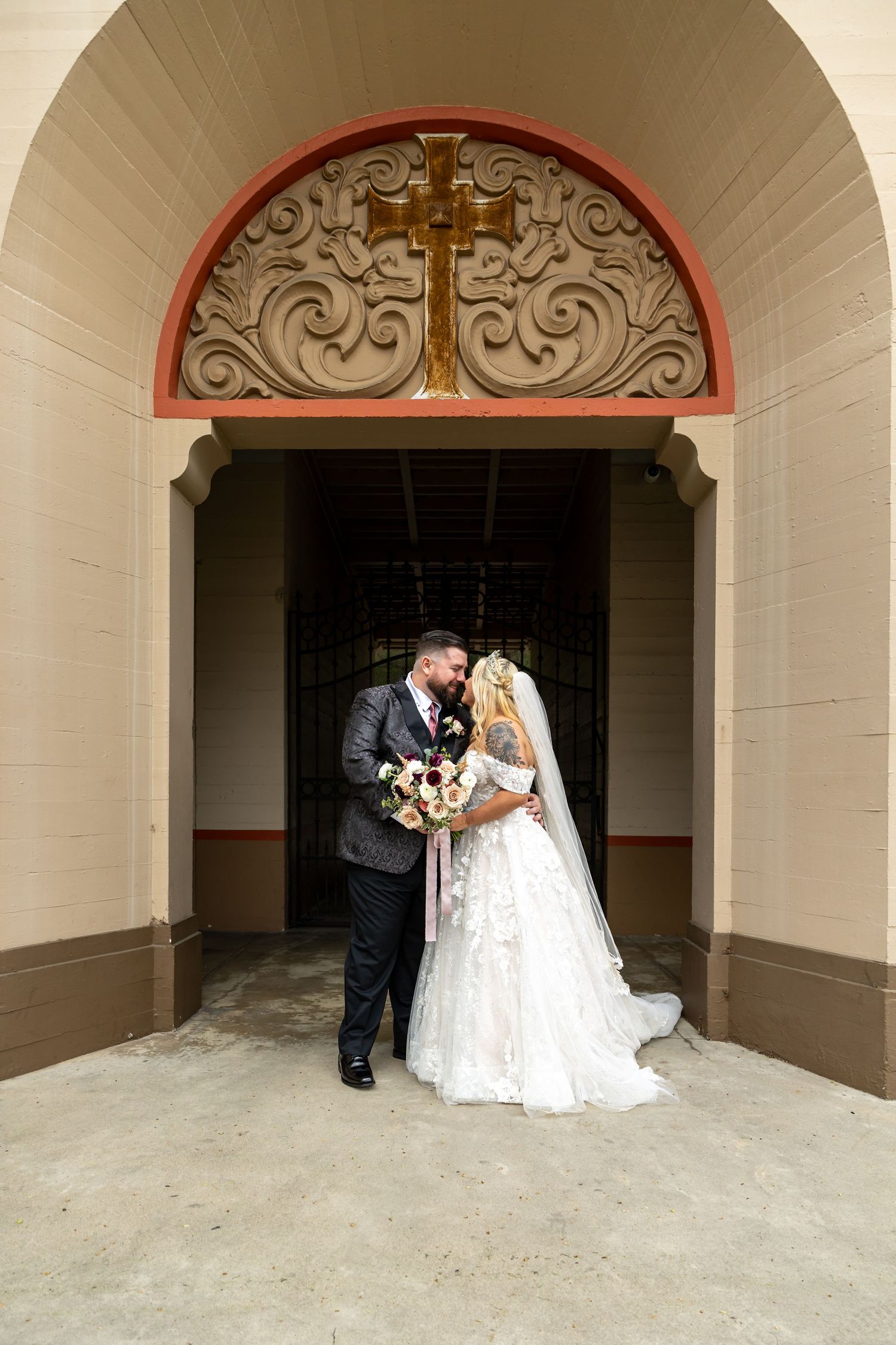 Wedding couple posing under arched church entrance with decorative cross detail in Santa Ana.