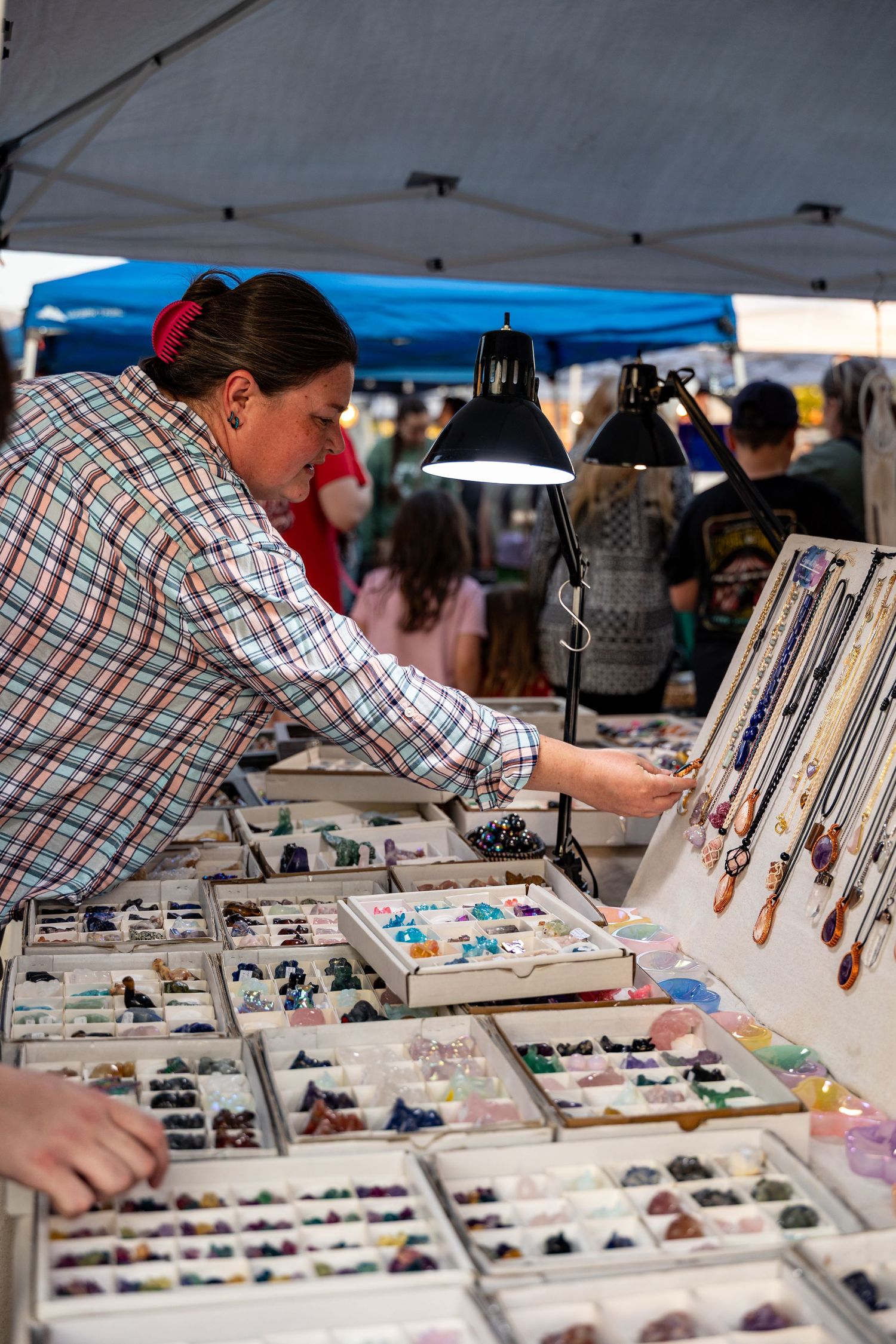 A vendor displays and arranges colorful jewelry pieces at an outdoor market stall under a blue tent canopy.