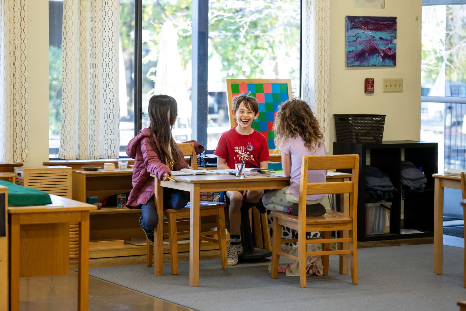 Students study and work at wooden tables in a library study area with tall windows letting in natural light.