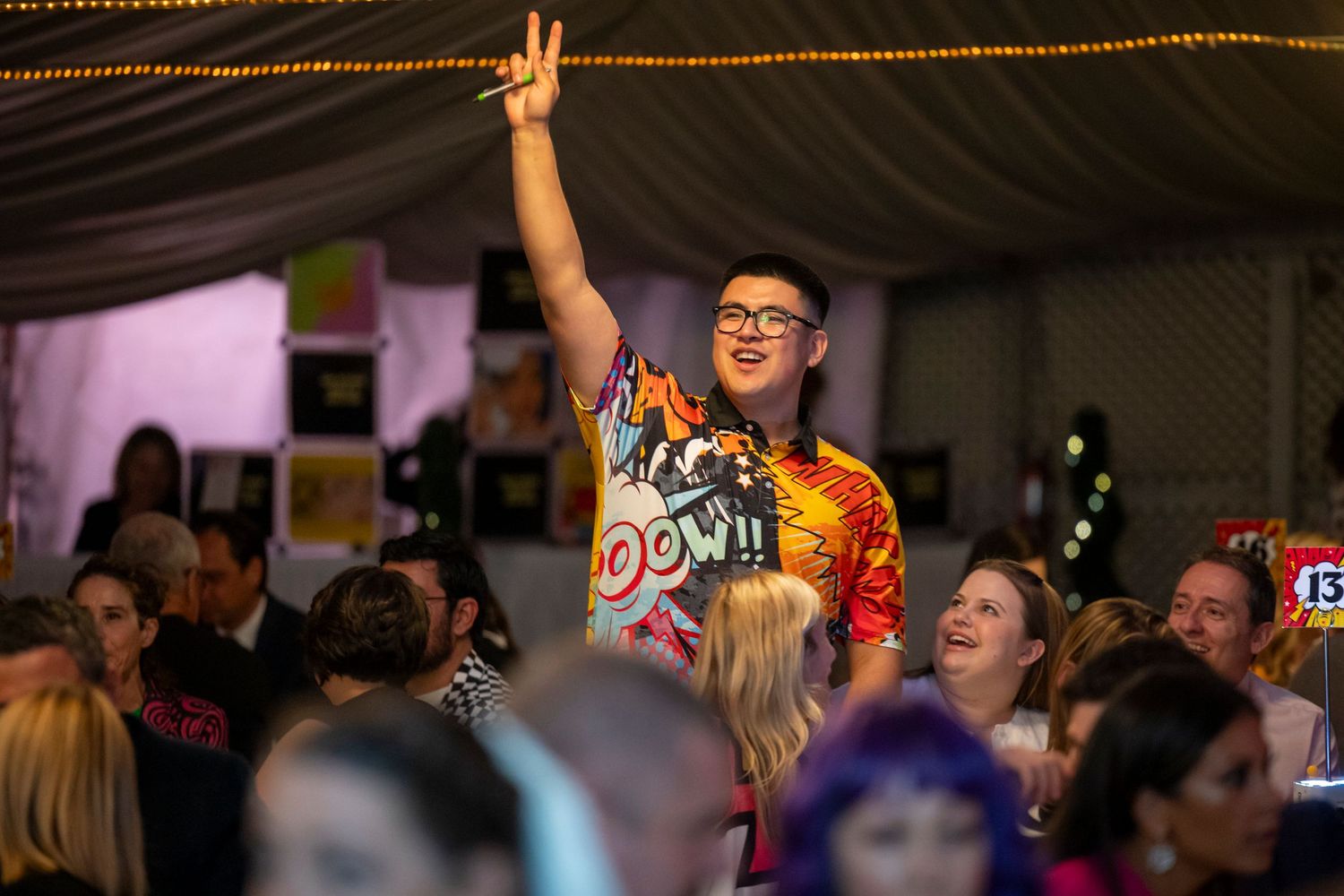 Large crowd enjoying a concert event with festive lighting and a raised V-sign hand gesture visible among the audience.