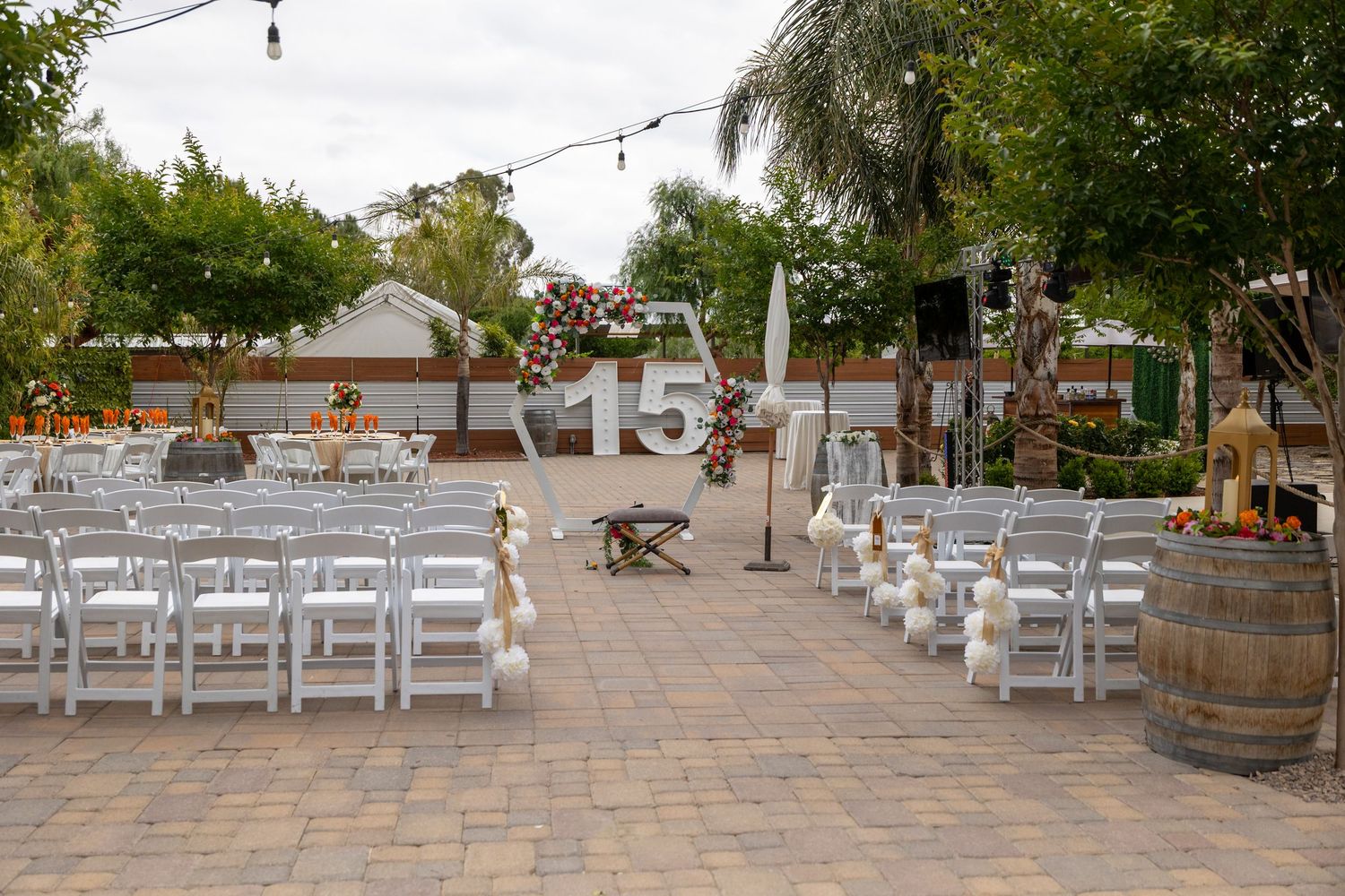 An outdoor wedding ceremony setup with white chairs and rustic wine barrels on a stone patio under string lights.